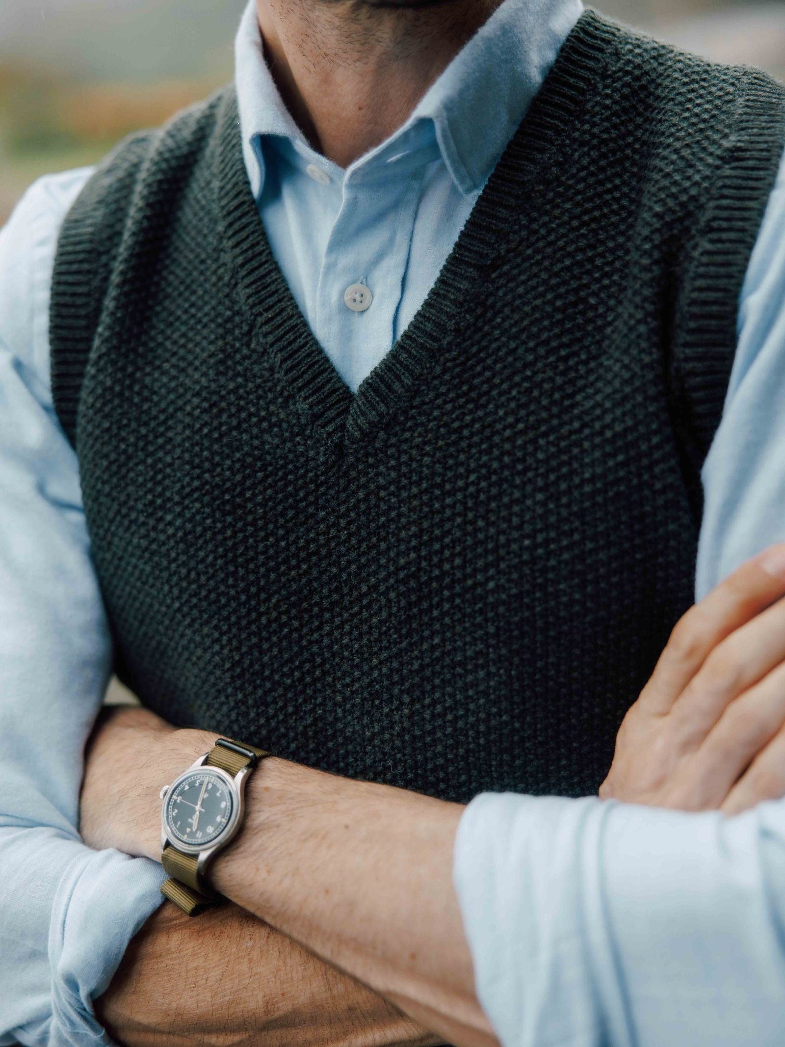 A person in a light blue shirt and Campbell's of Beauly Moss Stitch Knitted Slipover, made in the UK, stands with arms crossed, showing a fabric-strap wristwatch on their left wrist. The face is out of frame.