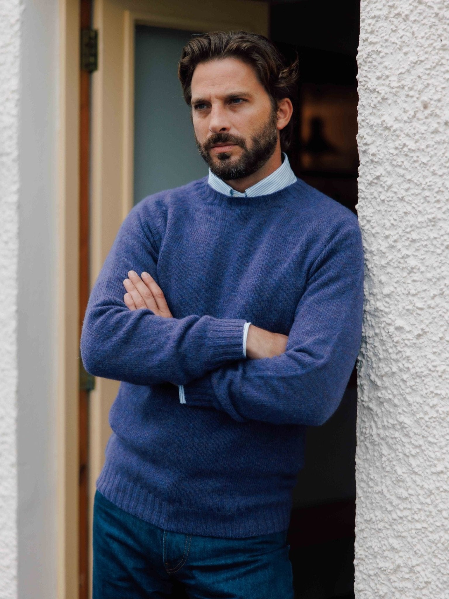 A man with dark hair and a beard, wearing Campbell's of Beauly Shetland Jumper over a collared shirt and blue jeans, stands with arms crossed against a white textured wall, gazing thoughtfully into the distance.