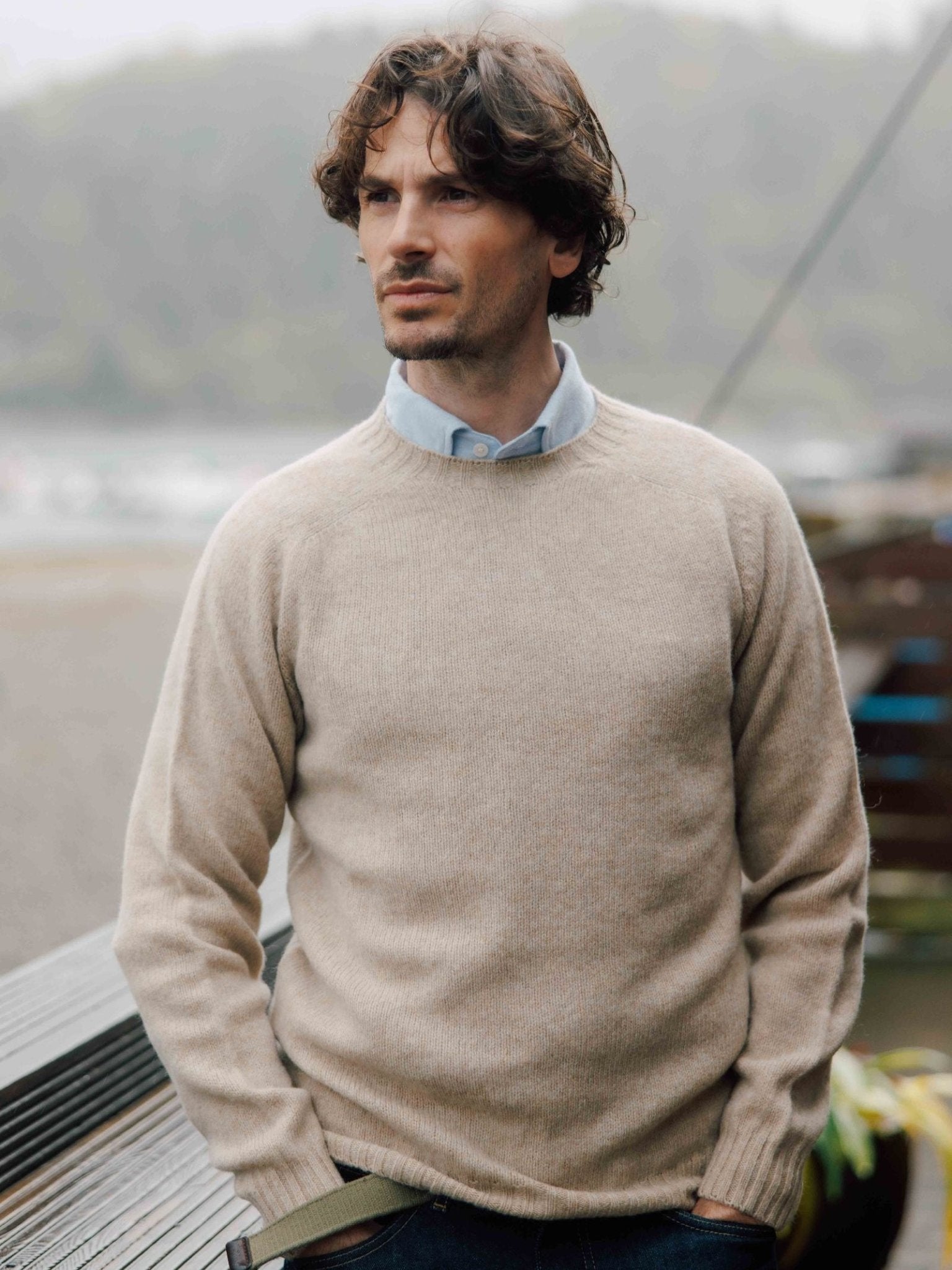 A man with wavy brown hair wears a beige Shetland Jumper by Campbell's of Beauly over a light blue collared shirt, leaning against a metallic railing outdoors with a blurred natural background.