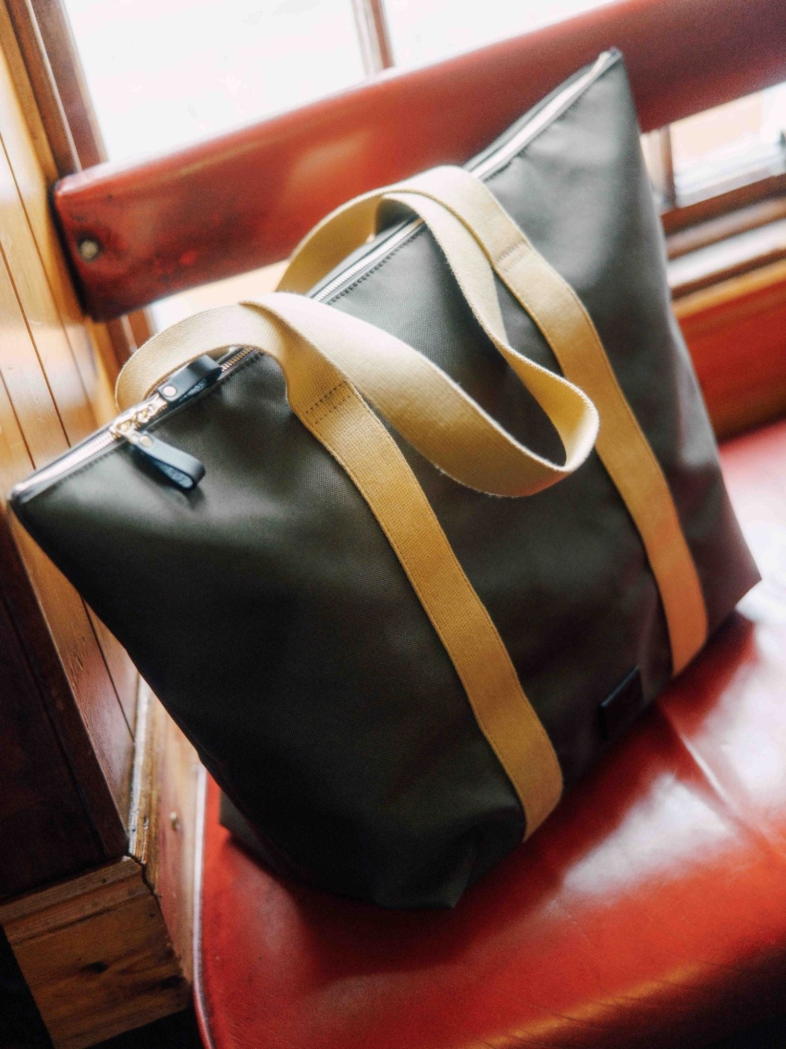 The Campbell's of Beauly Utility Medium Tote, a green canvas bag with tan straps, sits on a red cushioned seat by a wood-framed window—perfect for daily essentials.