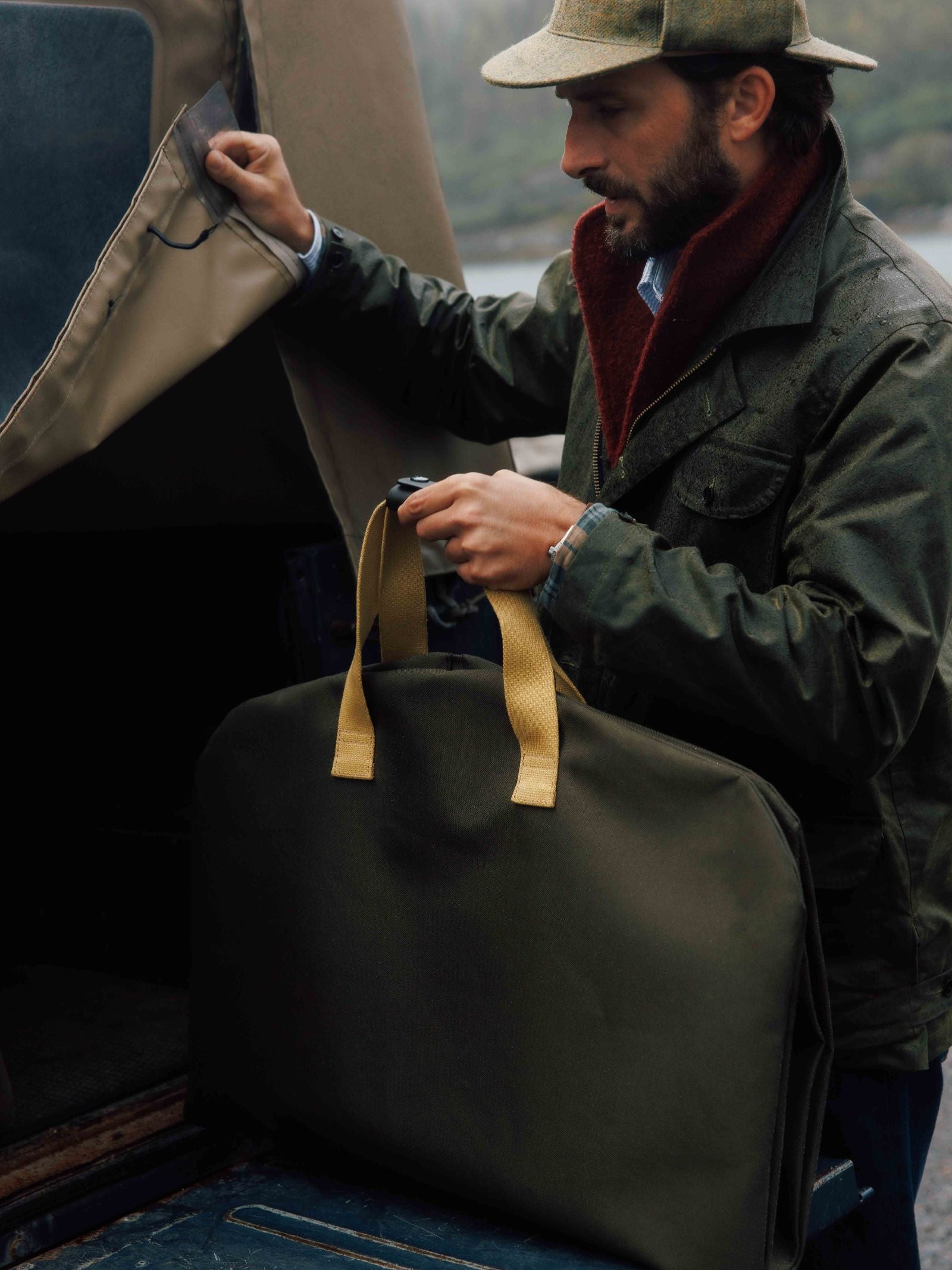 A man in a hat and jacket holds a large green Campbell's of Beauly Utility Suit Bag with tan handles, getting ready to place the garment bag into a vehicle with an open canvas cover for protection.