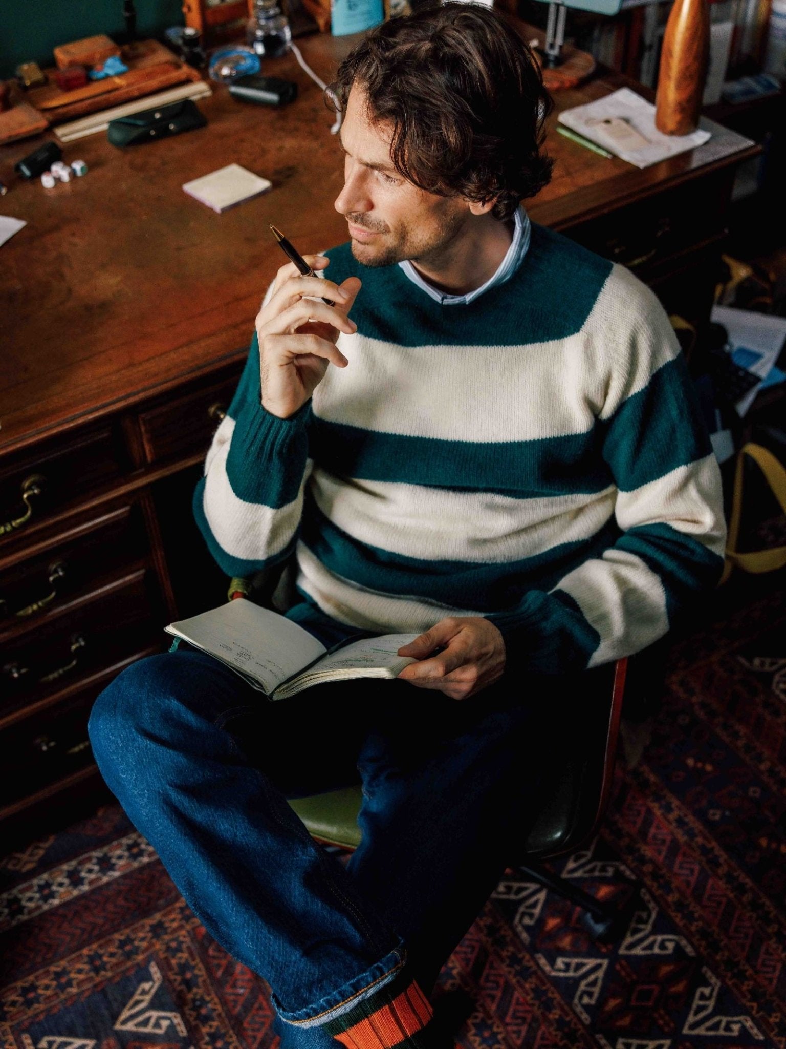 A man in a Campbell's of Beauly Shetland Stripe Crew Jumper sits cross-legged on a chair, smiling thoughtfully with a notebook and pen, at a wooden desk cluttered with books and papers on a patterned rug.