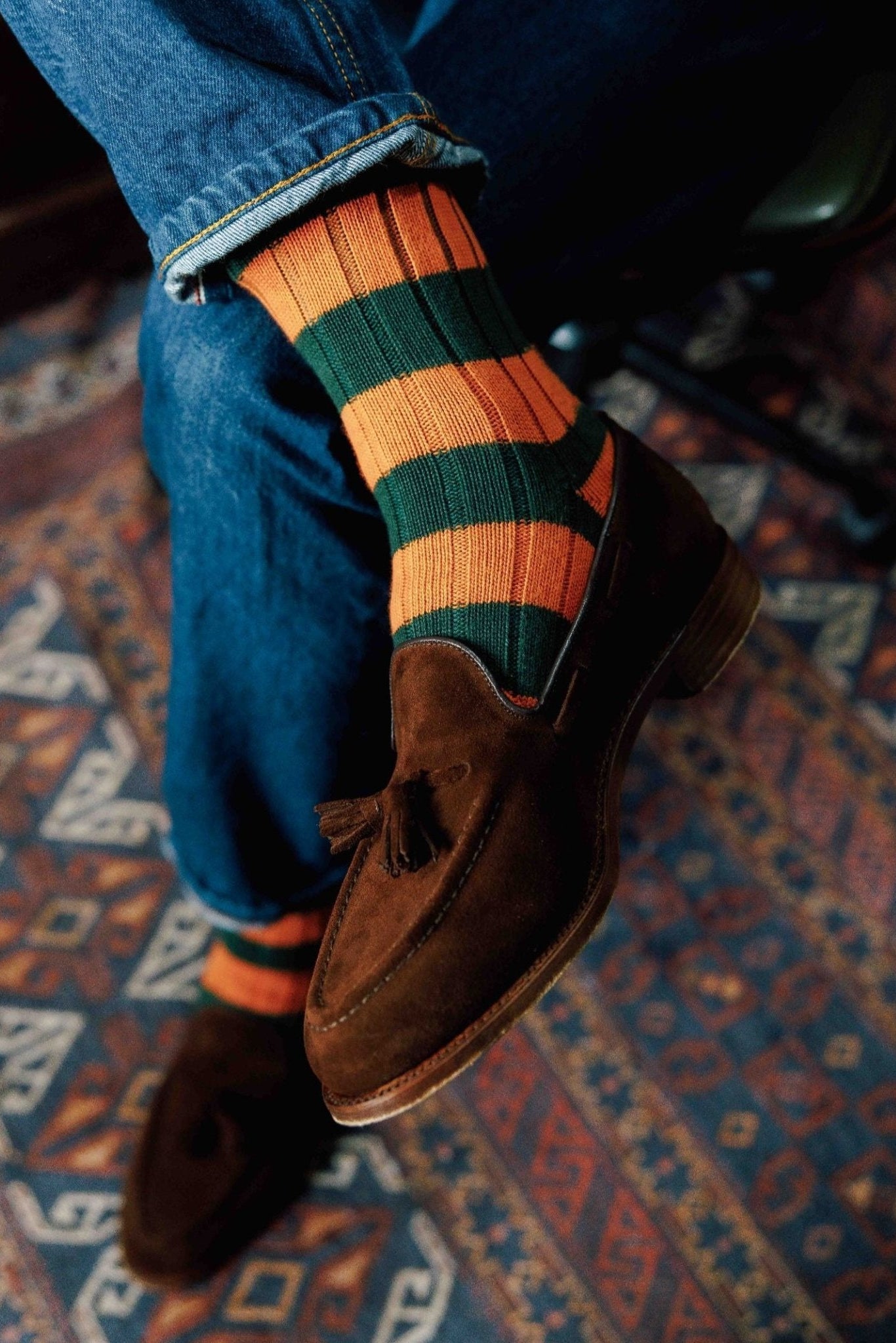 A person wearing blue jeans, brown suede tassel loafers, and Campbell's of Beauly Stripe Socks sits cross-legged on a patterned rug.