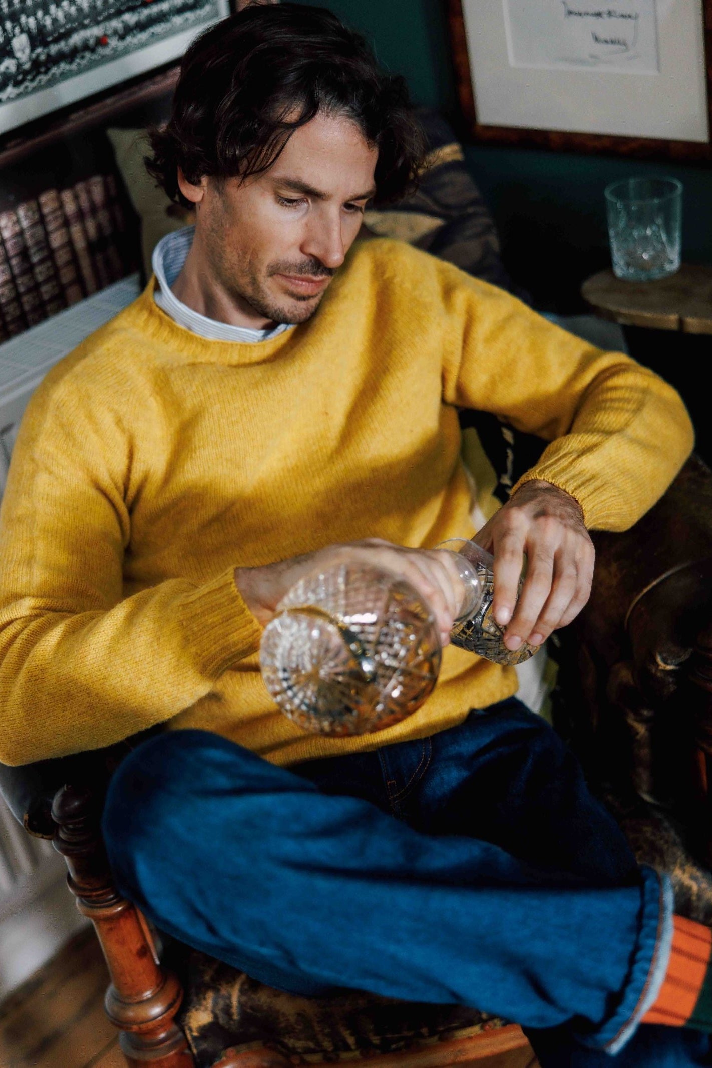 A man in a yellow Auld Stock Shetland Jumper by Campbell's of Beauly and blue jeans sits, pouring a drink from a crystal decanter into a glass, with another empty glass on the table nearby.
