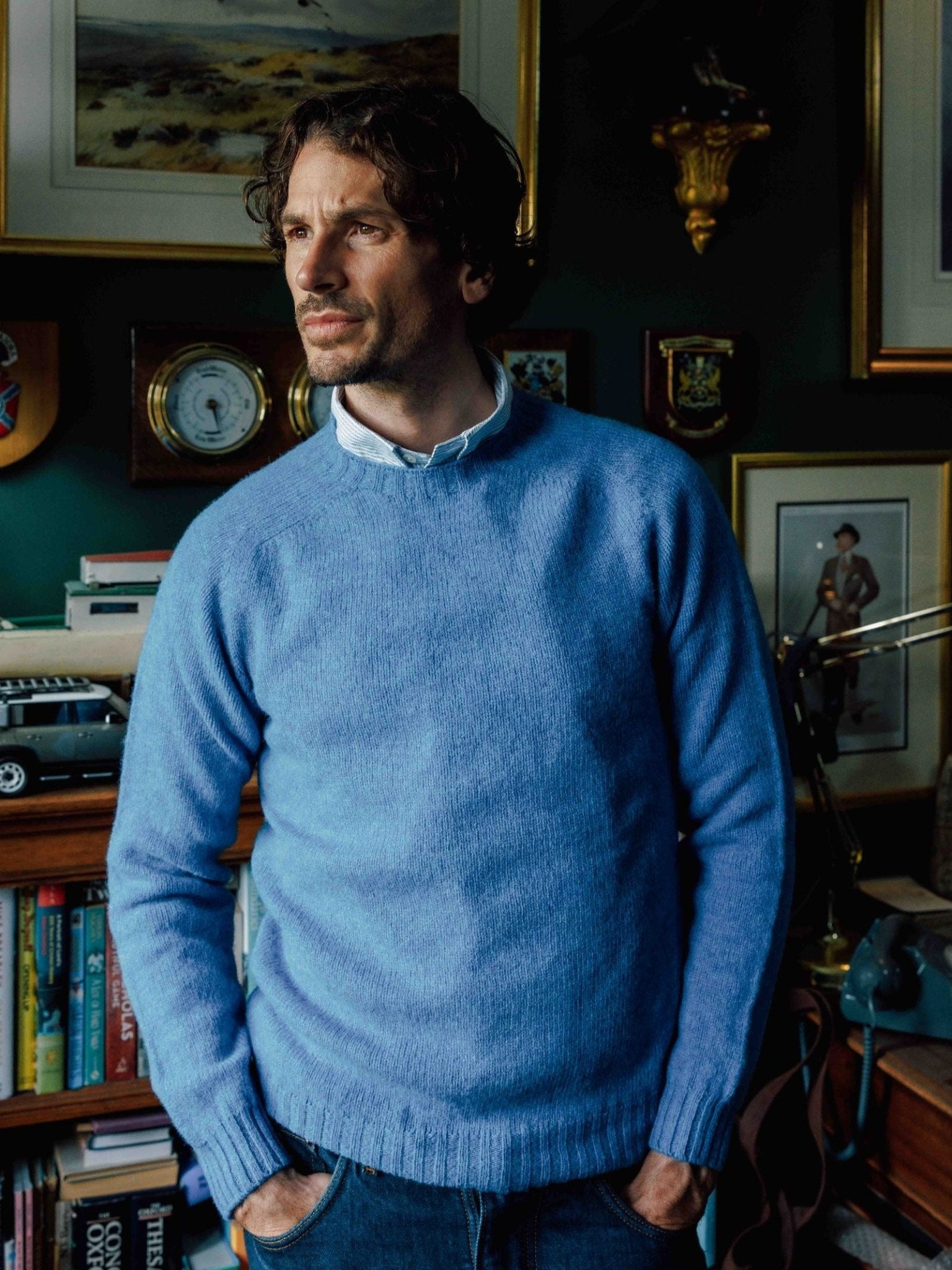 A man with wavy brown hair, in jeans and a light blue Campbell's of Beauly Shetland Jumper, stands indoors by a bookshelf and framed pictures, gazing thoughtfully to the side.