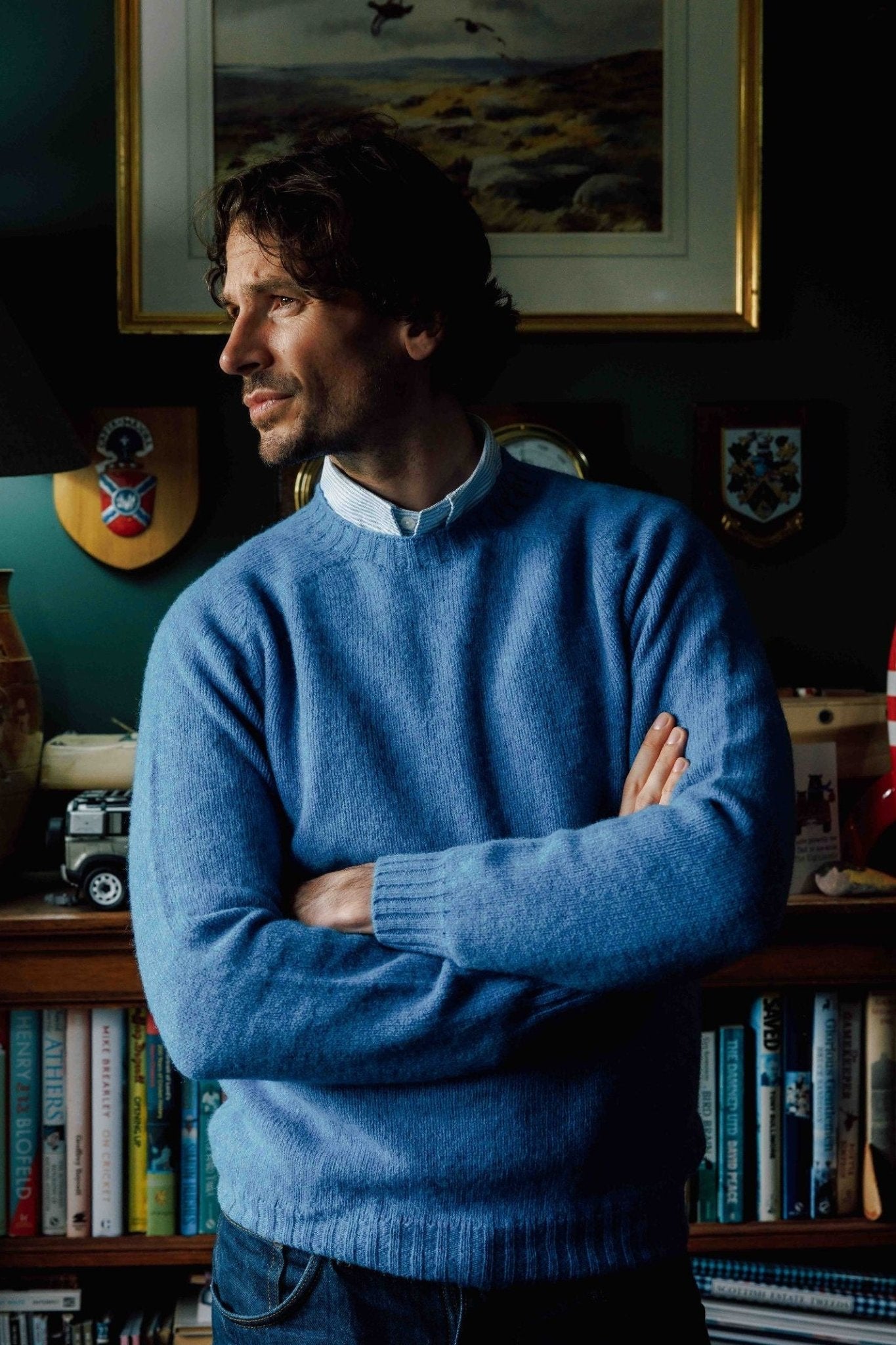 A man with wavy dark hair stands indoors, arms crossed and looking to the side, wearing a blue Campbell's of Beauly Shetland Jumper. Behind him are bookshelves, a lamp, framed art, and a red-and-white rocket model.