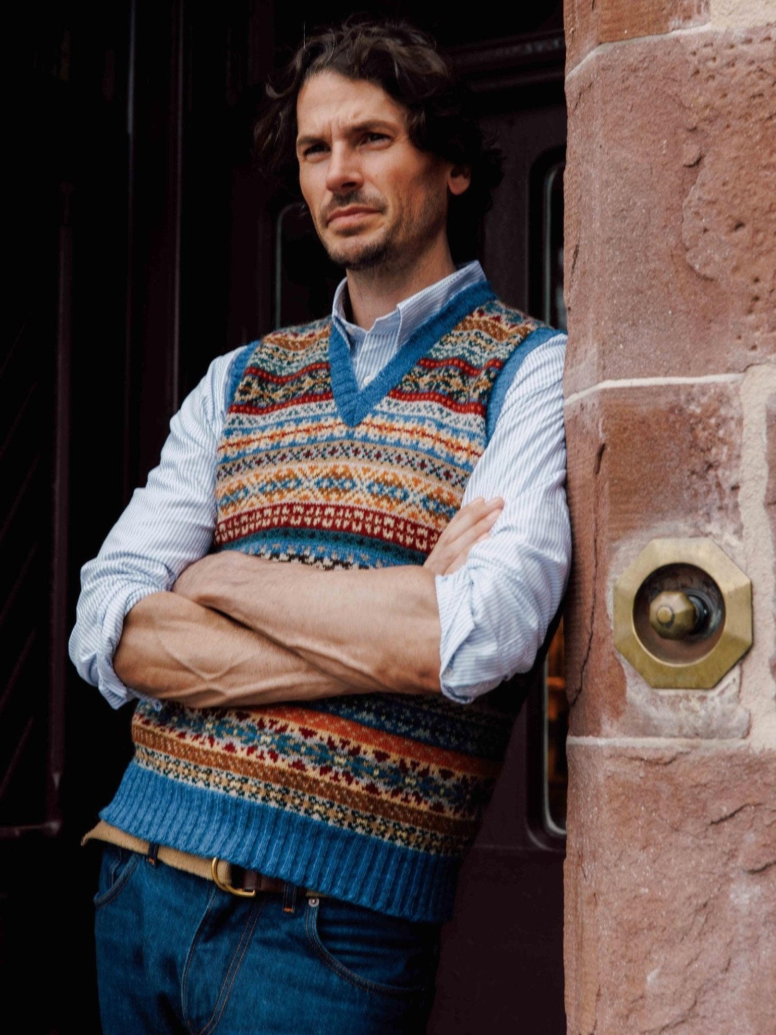 A man with wavy hair, arms crossed, wears the Campbell's of Beauly Fairisle Slipover—a colorful Shetland wool vest—over a striped shirt and jeans, leaning against a stone wall near a dark doorway.