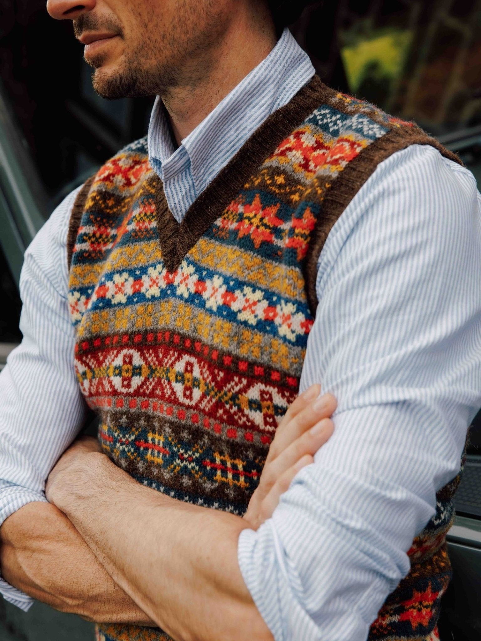 A man with folded arms wears a blue and white striped shirt and the Campbell's of Beauly Fairisle Slipover, a sleeveless Shetland wool layer with colorful geometric patterns. His face is partially out of frame.