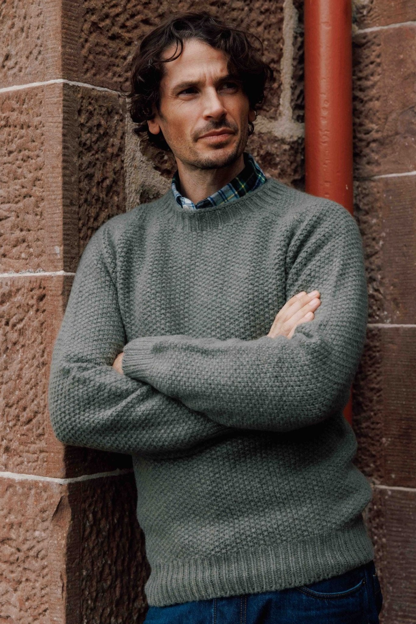 A man with curly brown hair, wearing a Campbell's of Beauly Moss Stitch Jumper over a green plaid shirt, stands with arms crossed against a stone wall, looking off to the side.