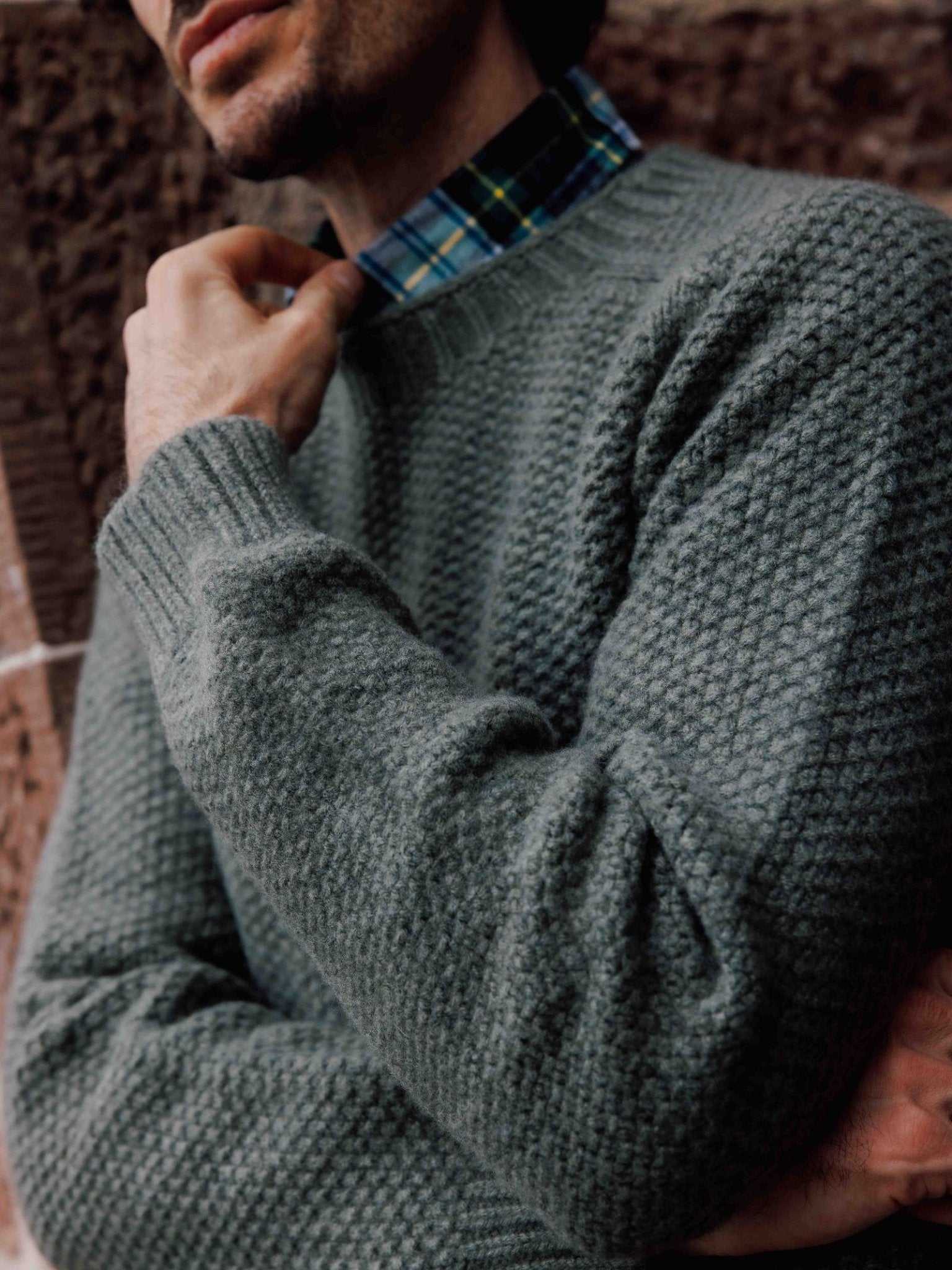 A person in a Campbell's of Beauly Moss Stitch Jumper, made in Scotland, layered over a blue and green plaid shirt, stands outside by a stone wall with hand near chin; their face is partly visible.