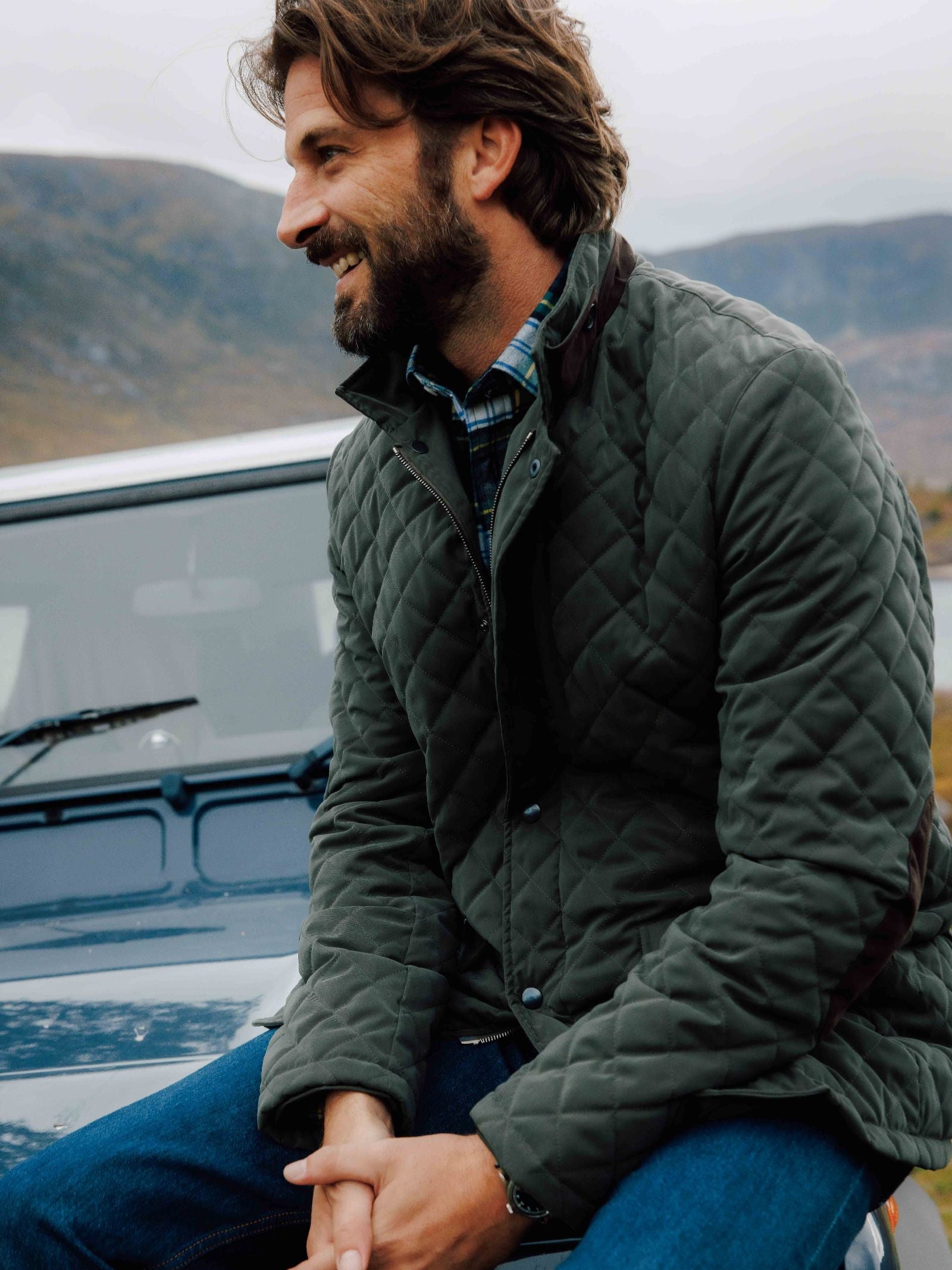 A man with brown hair and a beard, wearing the Tomich Jacket by Campbell's of Beauly and jeans, sits smiling on a blue car outdoors with hills and cloudy skies creating a Highland-inspired backdrop.