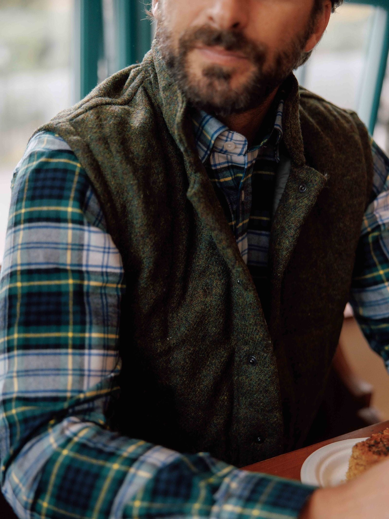 A man with a beard, partially out of frame, sits at a table near a window wearing the Campbell's of Beauly Auld Stock Harris Tweed Quilted Gilet over a blue and green plaid shirt. A plate is visible in the foreground.