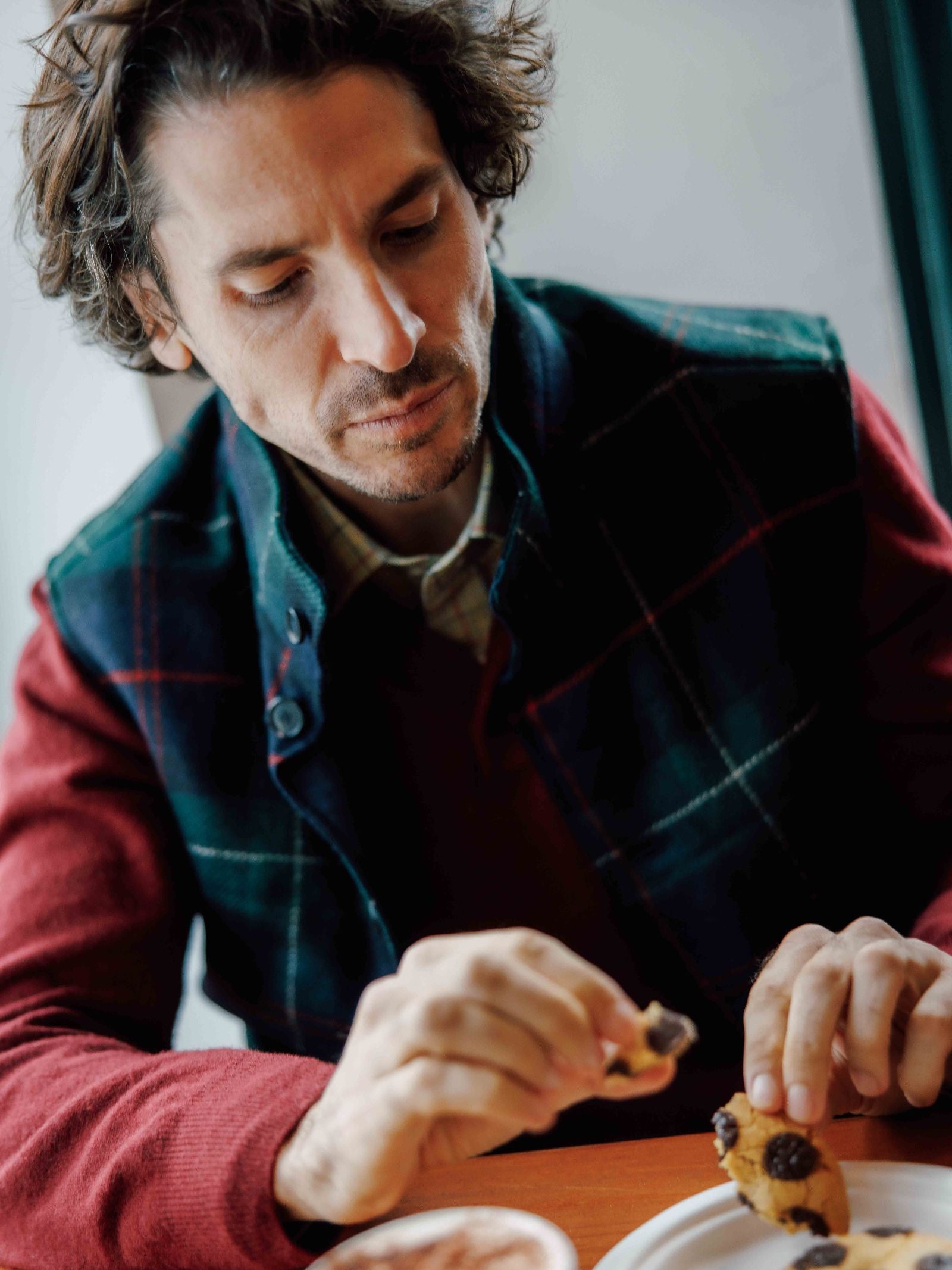A man with wavy brown hair wearing a Campbell's of Beauly Quilted Tweed Buttoned Gilet sits at a table, looking down as he prepares to eat a chocolate chip cookie, with a cup of coffee in front of him.