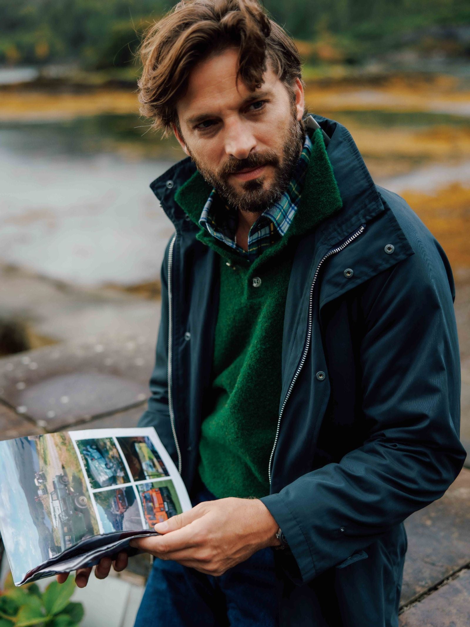 A man with wavy brown hair and a beard, wearing a Campbell's of Beauly Bouclé Polo Sweater over a plaid shirt and dark jacket, sits outdoors by the water, holding an open magazine and gazing slightly away from the camera.