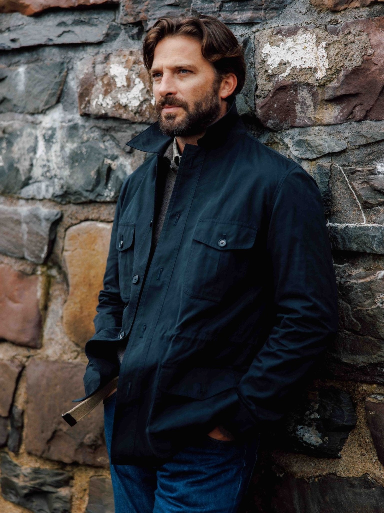 A man with brown hair and a beard stands outdoors against a stone wall, wearing the Campbell's of Beauly Aigas Field Jacket with his hands in his pockets, gazing thoughtfully into the distance.