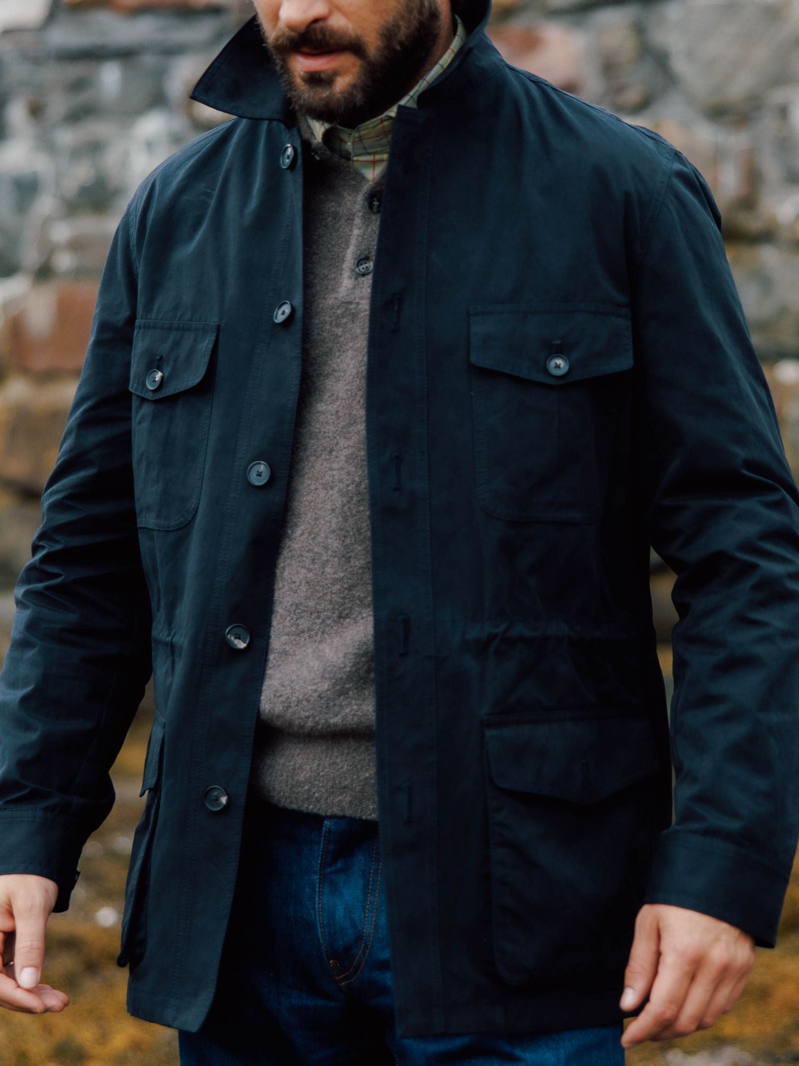 A man wearing the Campbell's of Beauly Aigas Field Jacket, crafted from water-repellent peach skin fabric, over a grey sweater and plaid shirt with blue jeans stands outdoors by a stone wall. His face is partially out of frame.