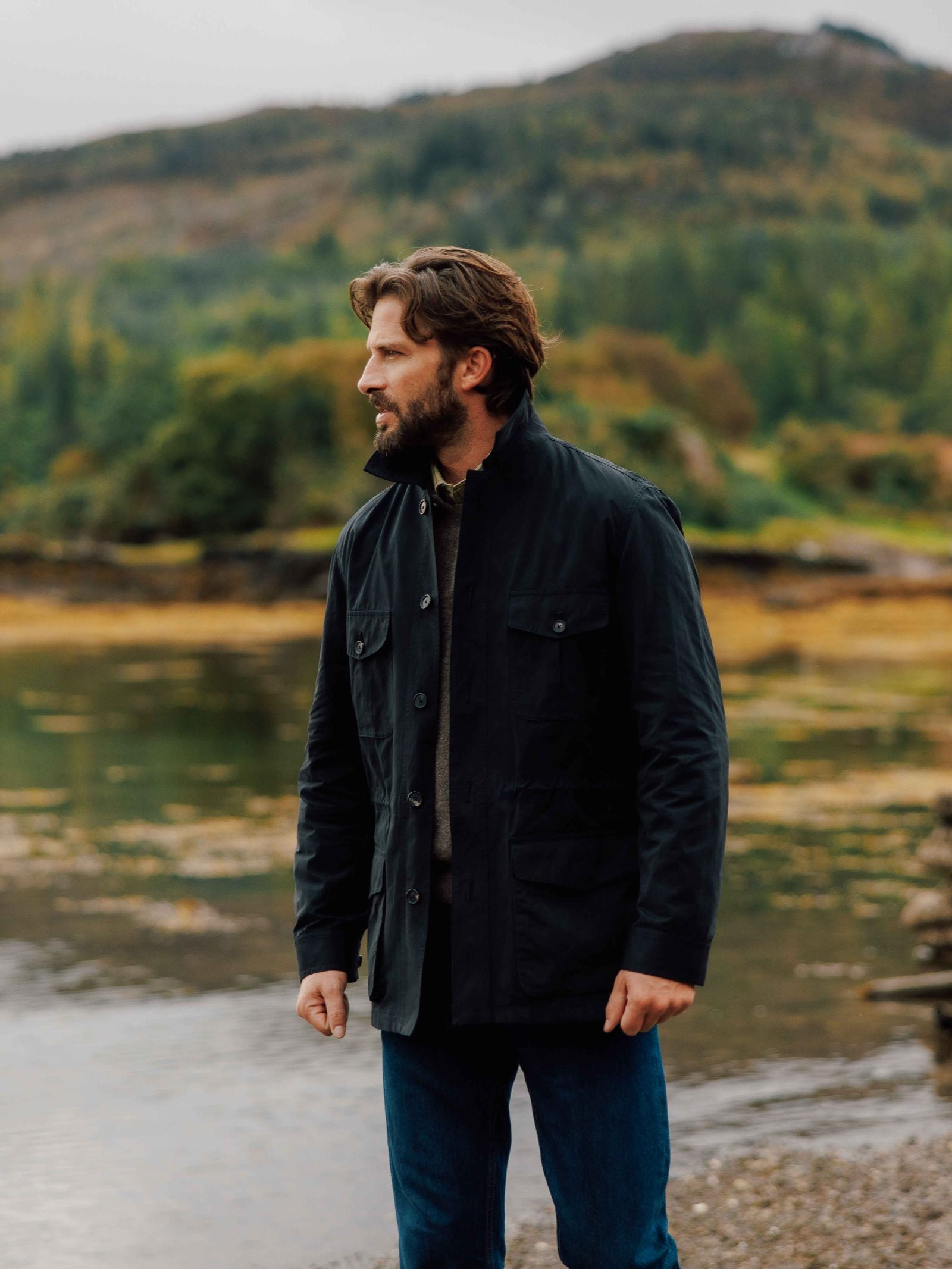 A bearded man in a water-repellent Campbell's of Beauly Aigas Field Jacket and blue jeans stands by a tranquil lakeshore with a forested hillside behind him, capturing a peaceful, outdoorsy vibe.