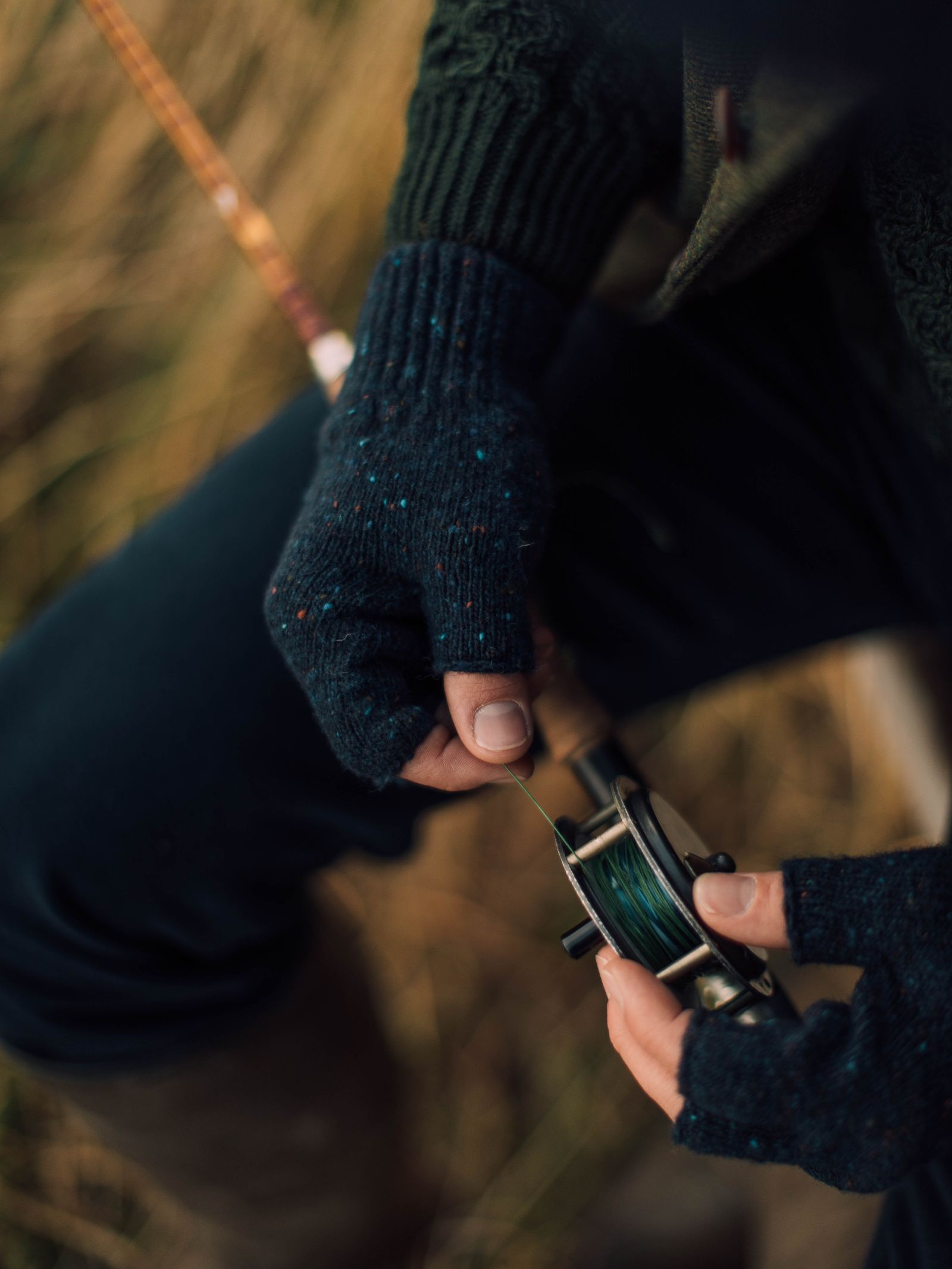 Wearing Campbells of Beauly Donegal Lambswool Half-Finger Gloves, a person adjusts a fishing reel outdoors, with grass blurred in the background.