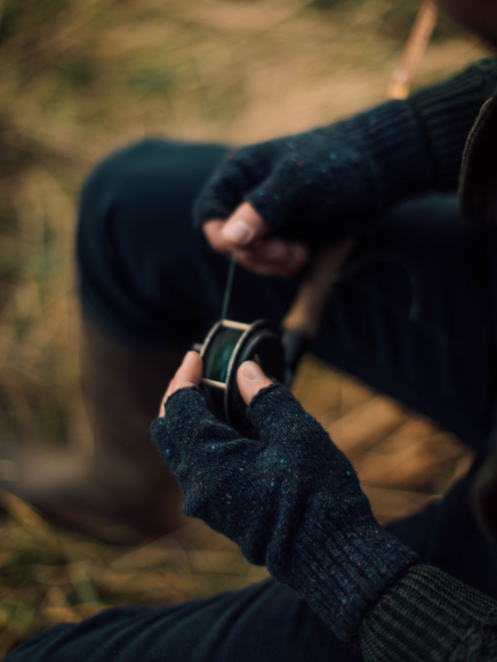 A person wearing Campbells of Beauly Donegal Lambswool Half-Finger Gloves winds fishing line onto a reel while sitting outdoors on grass or straw, with only their hands and lap visible in the image.