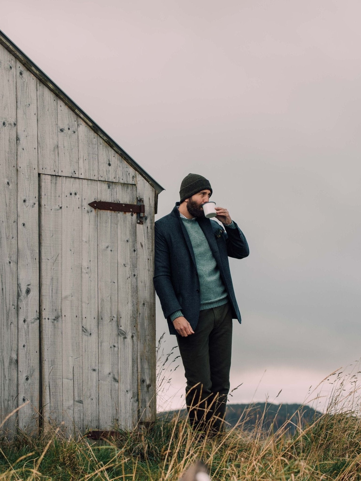A man in a Campbells of Beauly Glen Check Jacket, paired with a beanie and sweater, stands by a weathered wooden shed sipping from a white mug. Tall grass and an overcast sky create a calm, rustic outdoor atmosphere.