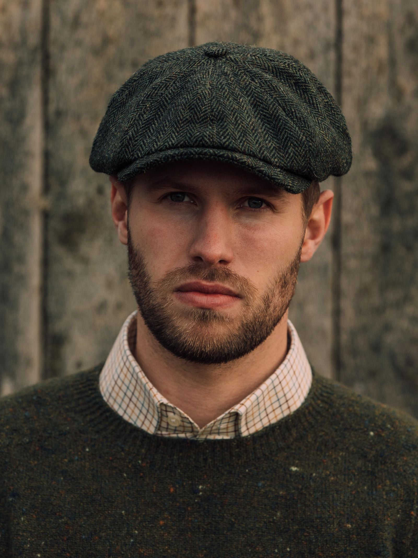 A man with a short beard and mustache wears the Farrar Cap by Campbells of Beauly, paired with a green sweater and checked shirt, standing before a weathered wooden wall and looking directly at the camera with a neutral expression.