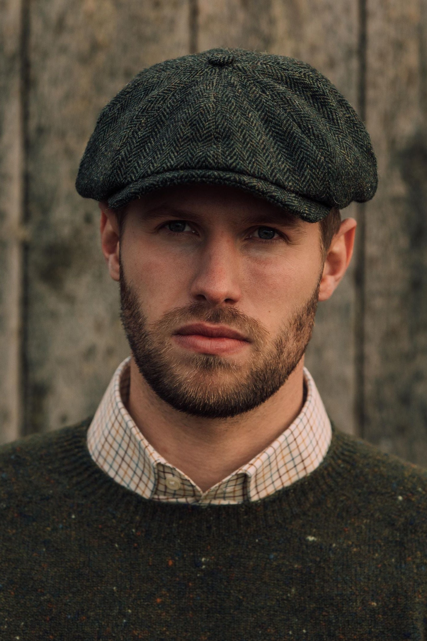 A man with a short beard and mustache wears the Farrar Cap by Campbells of Beauly, paired with a green sweater and checked shirt, standing before a weathered wooden wall and looking directly at the camera with a neutral expression.