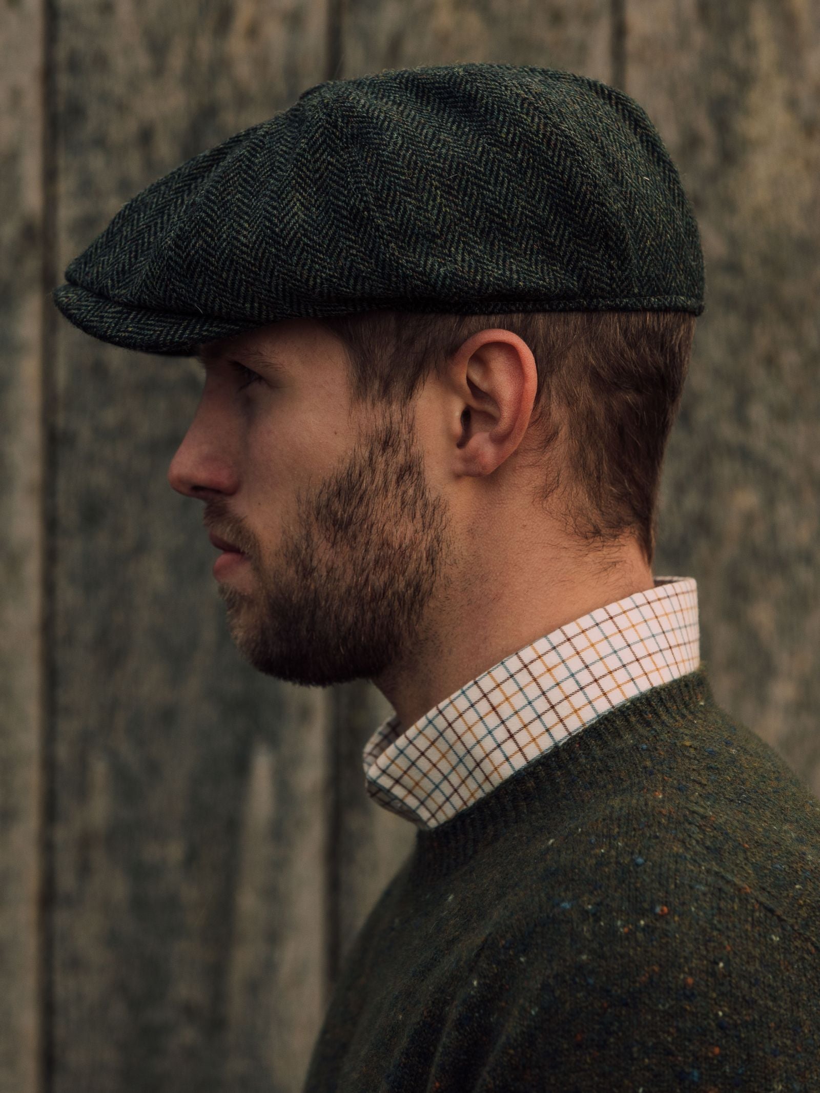 A bearded man in profile wears the Campbells of Beauly Farrar Cap, a green sweater, and a white plaid shirt, standing before a wooden backdrop reminiscent of British country heritage.