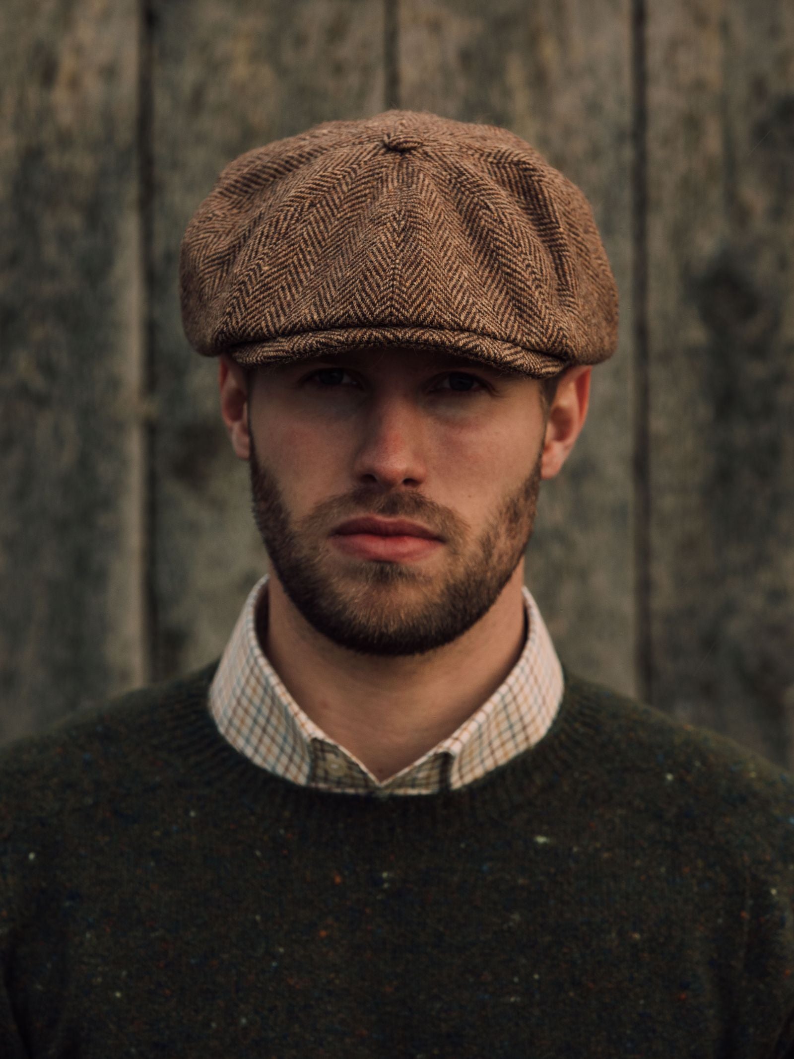 A man with a trimmed beard wears the Campbells of Beauly Farrar Cap, paired with a checkered shirt and dark green sweater, standing against a weathered wooden background and looking directly at the camera with a neutral expression.