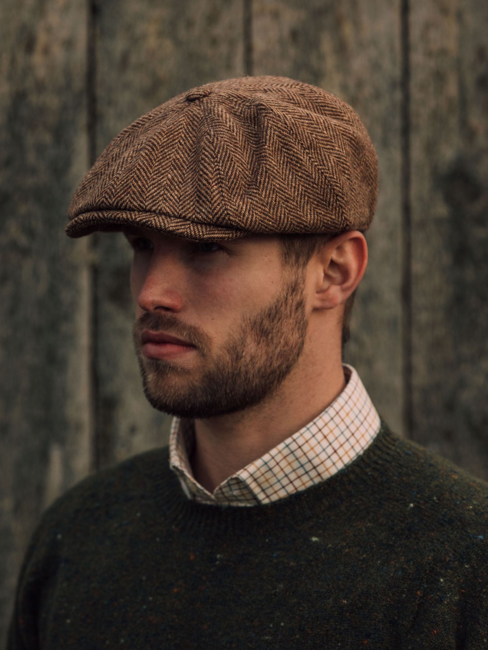 Wearing a Campbells of Beauly Farrar Cap, a bearded man in a green sweater and checked shirt stands before weathered wood, embodying classic British country heritage style.