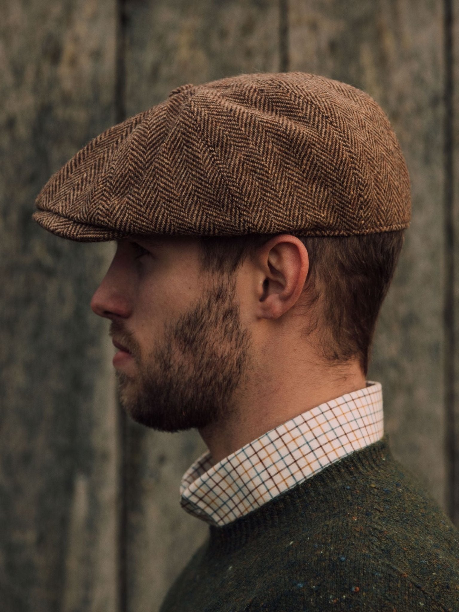 A bearded man in profile wears the Farrar Cap by Campbells of Beauly, paired with a green sweater and checkered shirt, standing before a weathered wooden wall that reflects classic British country heritage.
