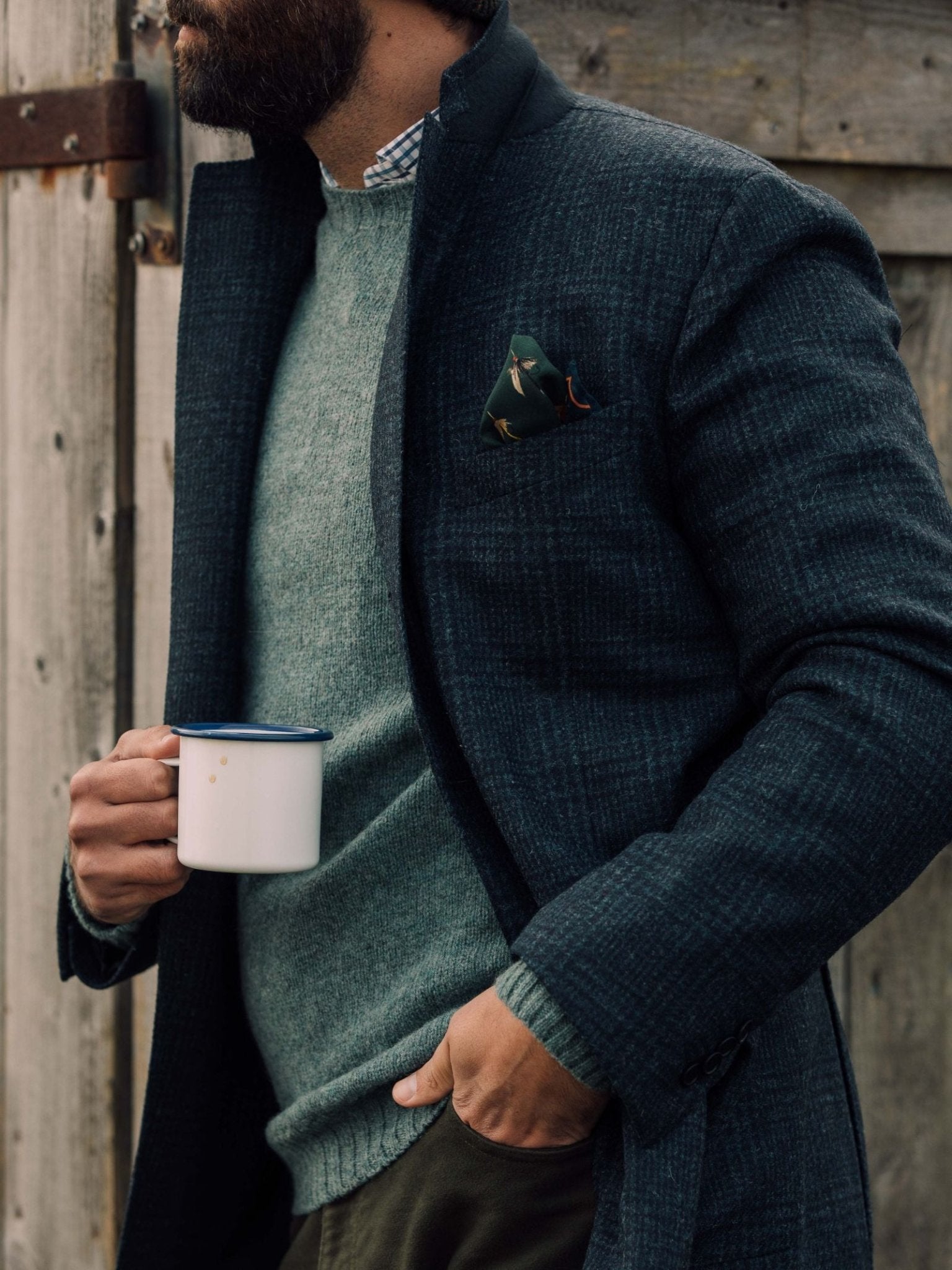 A bearded man stands outdoors by a wooden fence, wearing Campbells of Beauly Glen Check Jacket over a green sweater. A patterned pocket square peeks from his jacket as he holds a white mug with a blue rim.