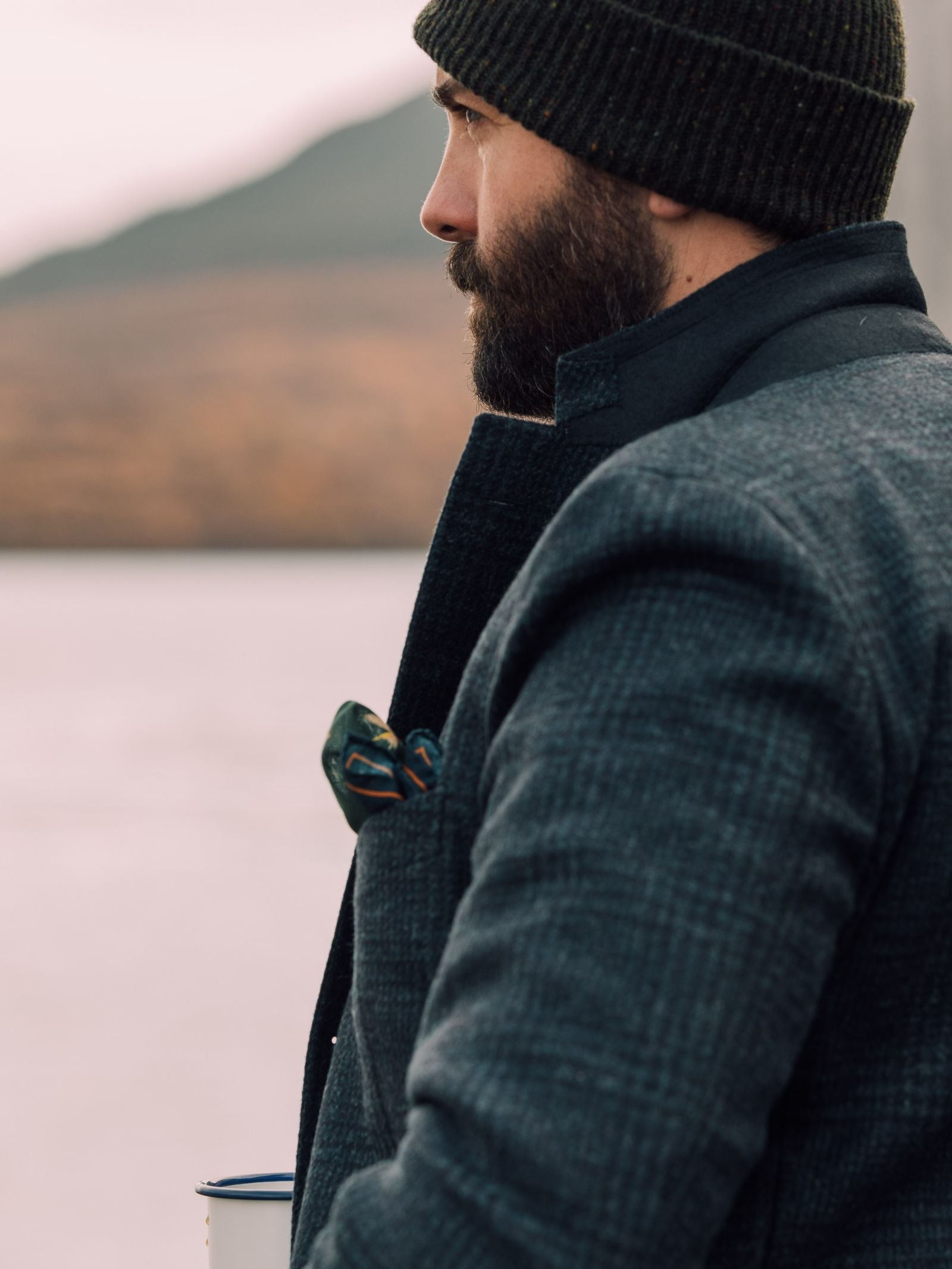 A bearded man in a Glen Check Jacket by Campbells of Beauly stands outdoors, holding a white mug and gazing into the distance, the blurred mountains behind him reflecting classic tailoring style.