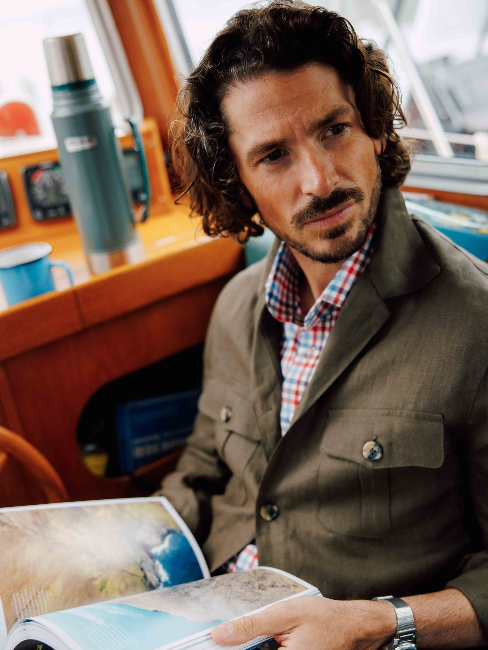 A man with curly brown hair and a beard wears the Campbells of Beauly Linen Safari Jacket over a checked shirt as he sits indoors, holding an open magazine, with a thermos and blue mug on the wooden surface behind him.
