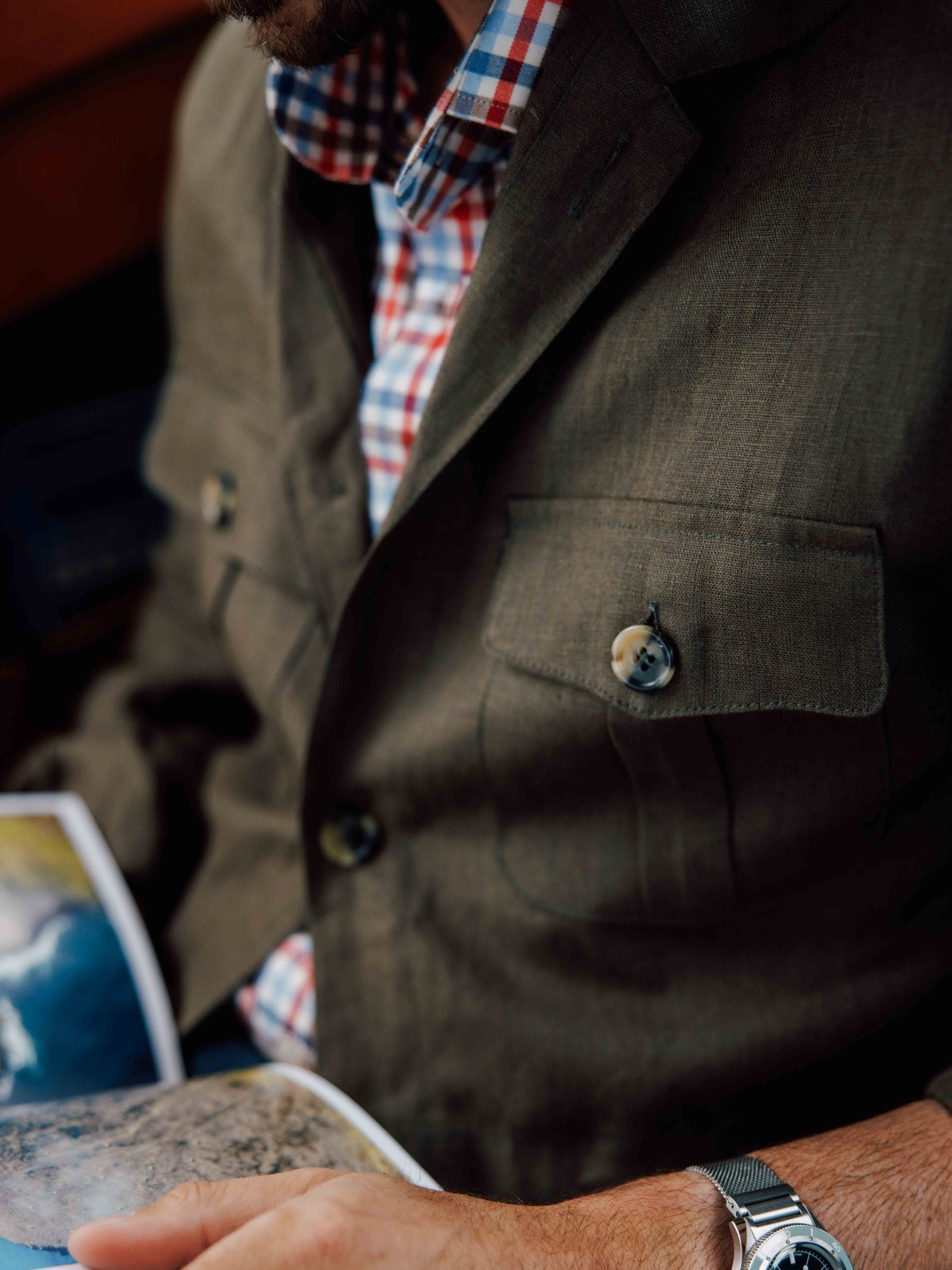 A man wears the Campbell’s of Beauly Linen Safari Jacket over a red-and-blue plaid shirt, holding an open magazine. The shot focuses on his torso and silver wristwatch, highlighting this relaxed layered look.