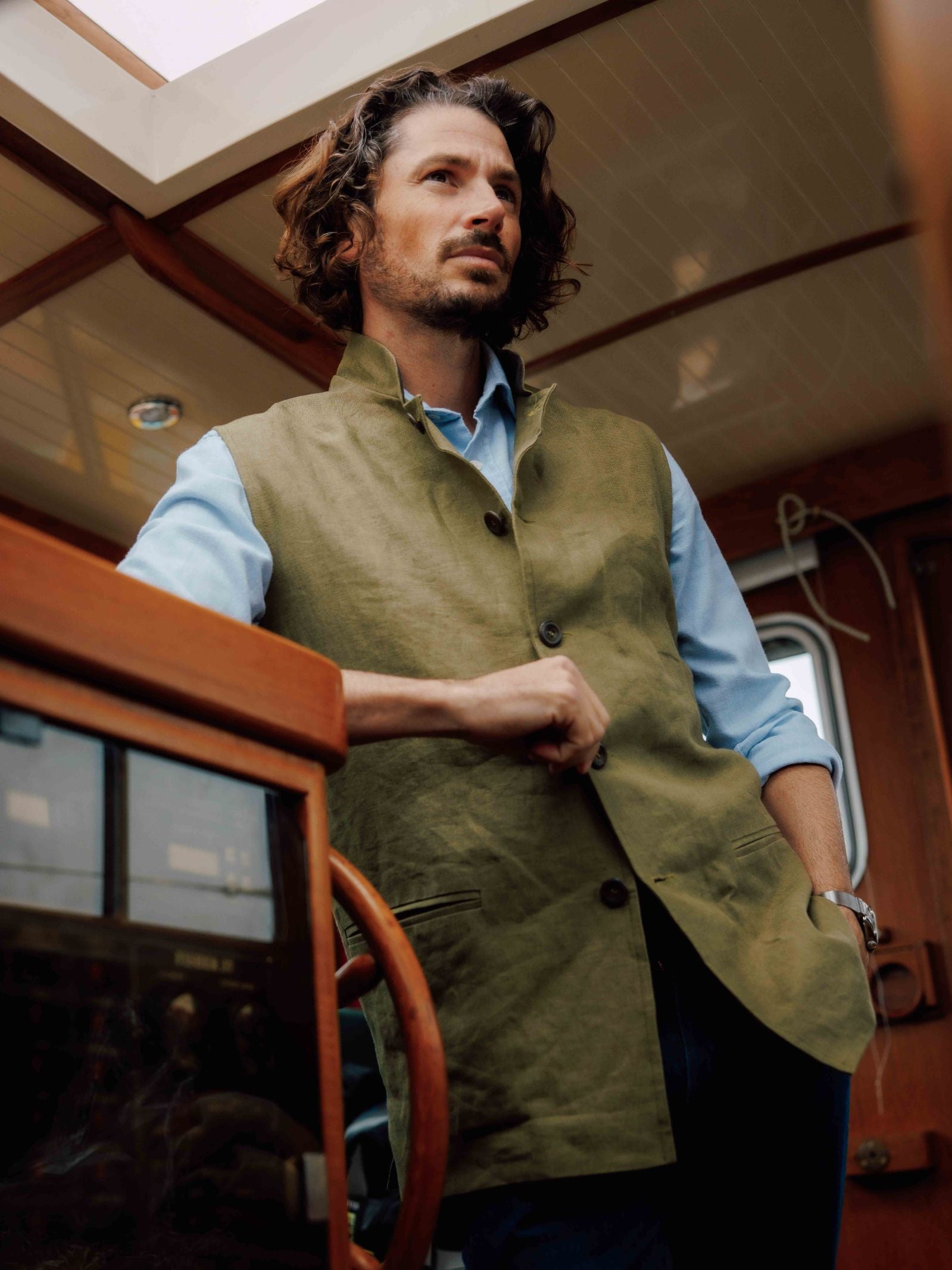 A man with wavy hair and a beard stands in a wooden boat, wearing a light blue shirt and the Linen Nehru Gilet by Campbell’s of Beauly—his ideal summer layer—as he rests one hand on the wheel, gazing thoughtfully into the distance.