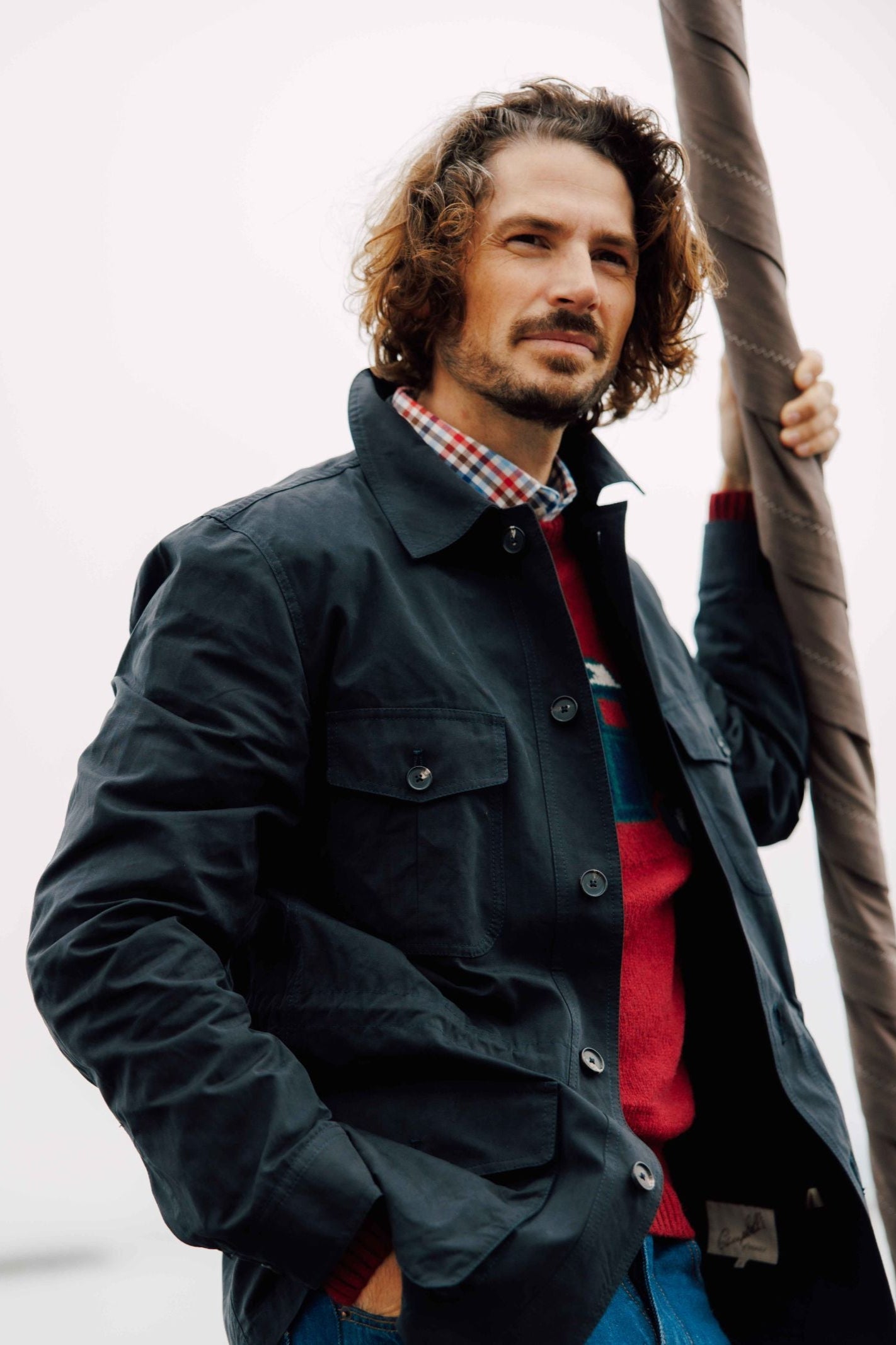 A man with wavy brown hair and a beard stands outdoors, confidently wearing Campbells of Beauly Aigas Field Jacket with a water-repellent finish over a red sweater and plaid shirt, holding a pole in one hand.