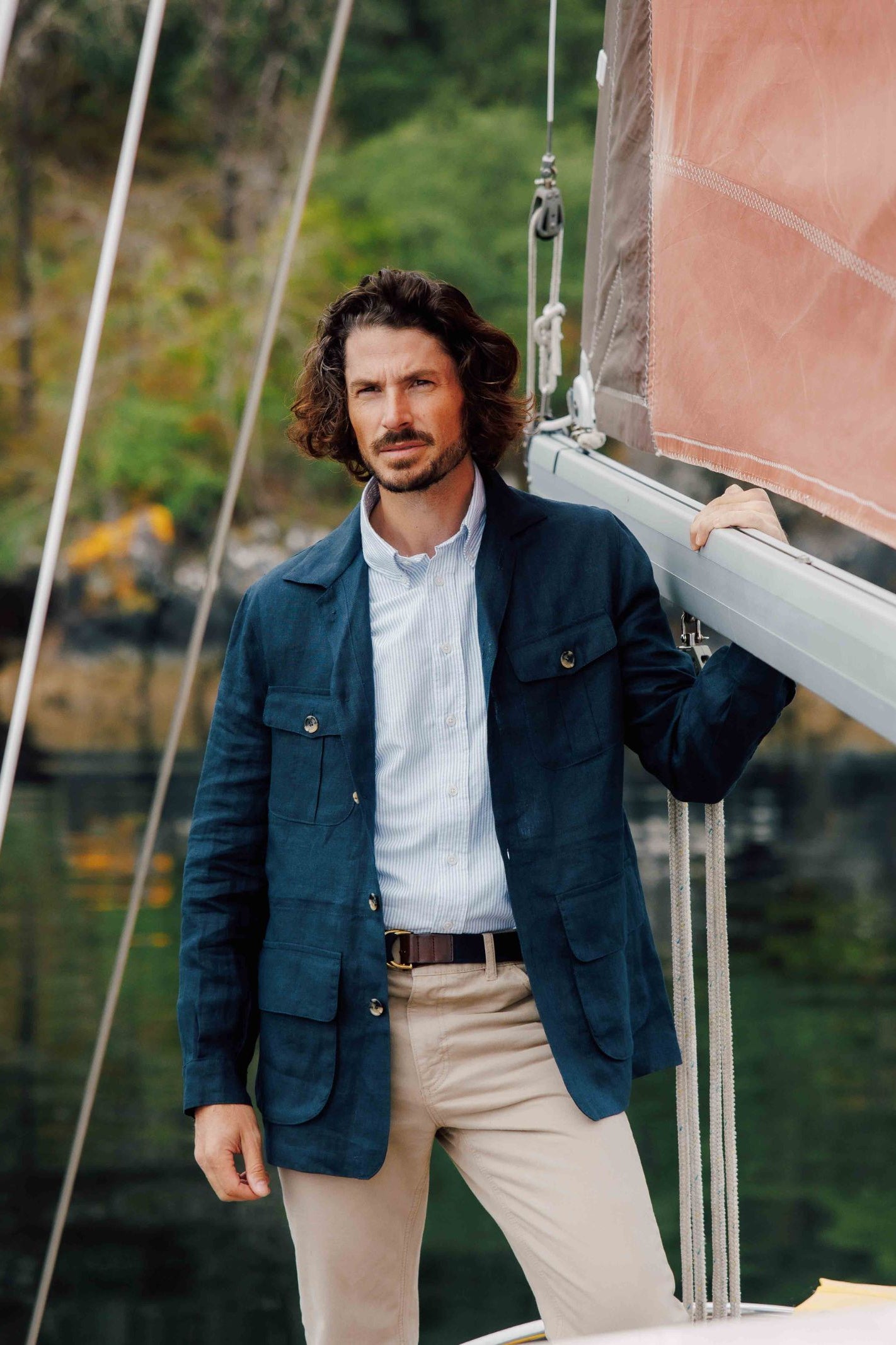 A man with wavy brown hair and a beard stands on a sailboat, wearing the Campbells of Beauly Linen Safari Jacket as a casual layer, one hand resting on the rigging. Green foliage and water form the background.