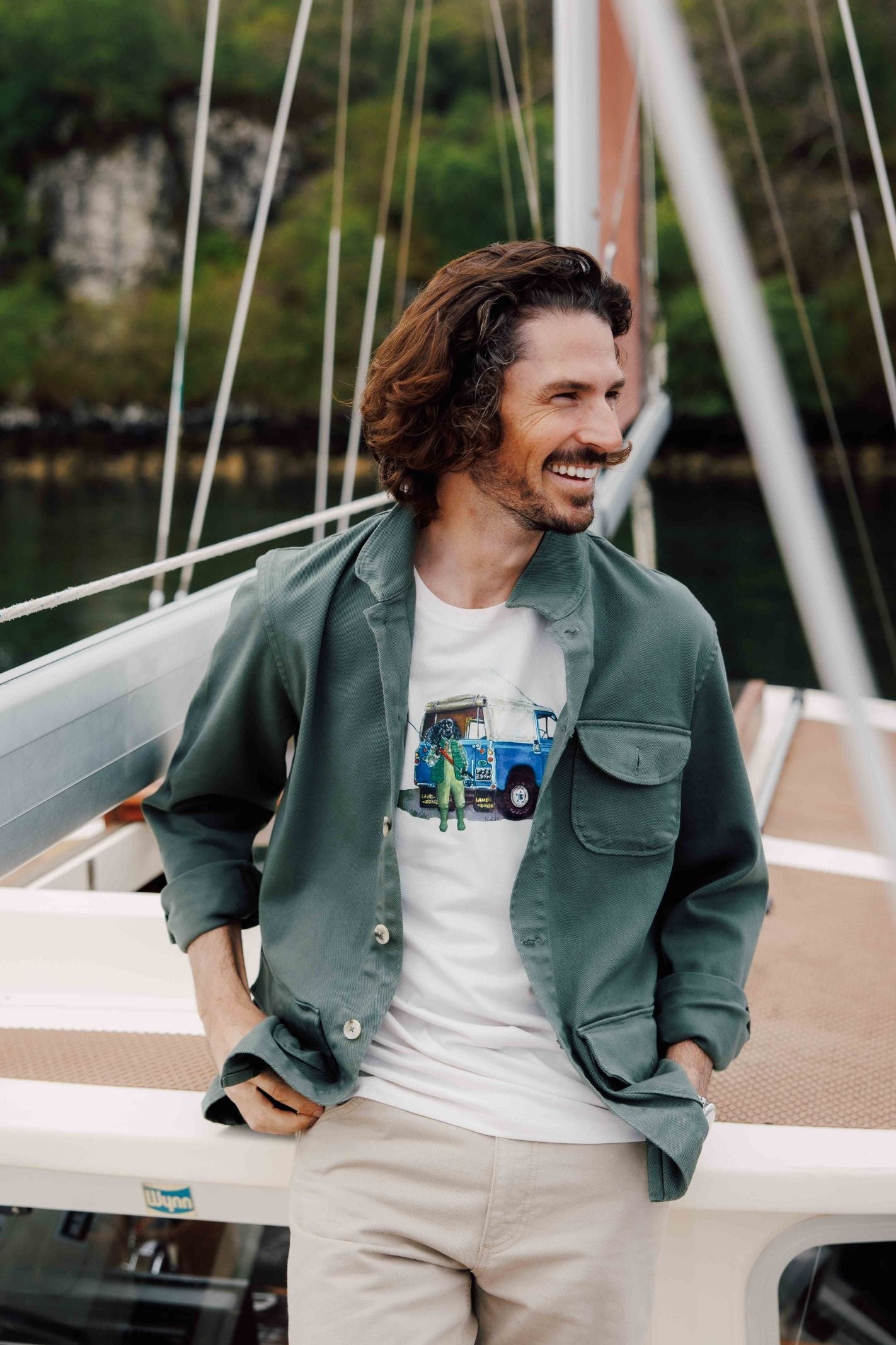 A man with wavy brown hair smiles on a sailboat, wearing the Campbells of Beauly Field Chore Jacket over a graphic T-shirt and beige pants, with water and greenery in the background.