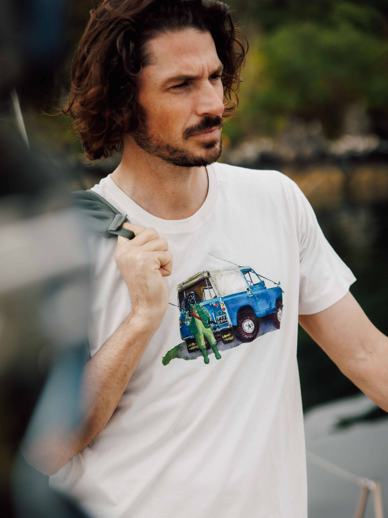 A man with wavy brown hair and a beard stands outdoors, carrying a bag over his shoulder. He is wearing the Campbells of Beauly Land Rover Cocker T-Shirt, which features an illustration of two people near a blue camper van.