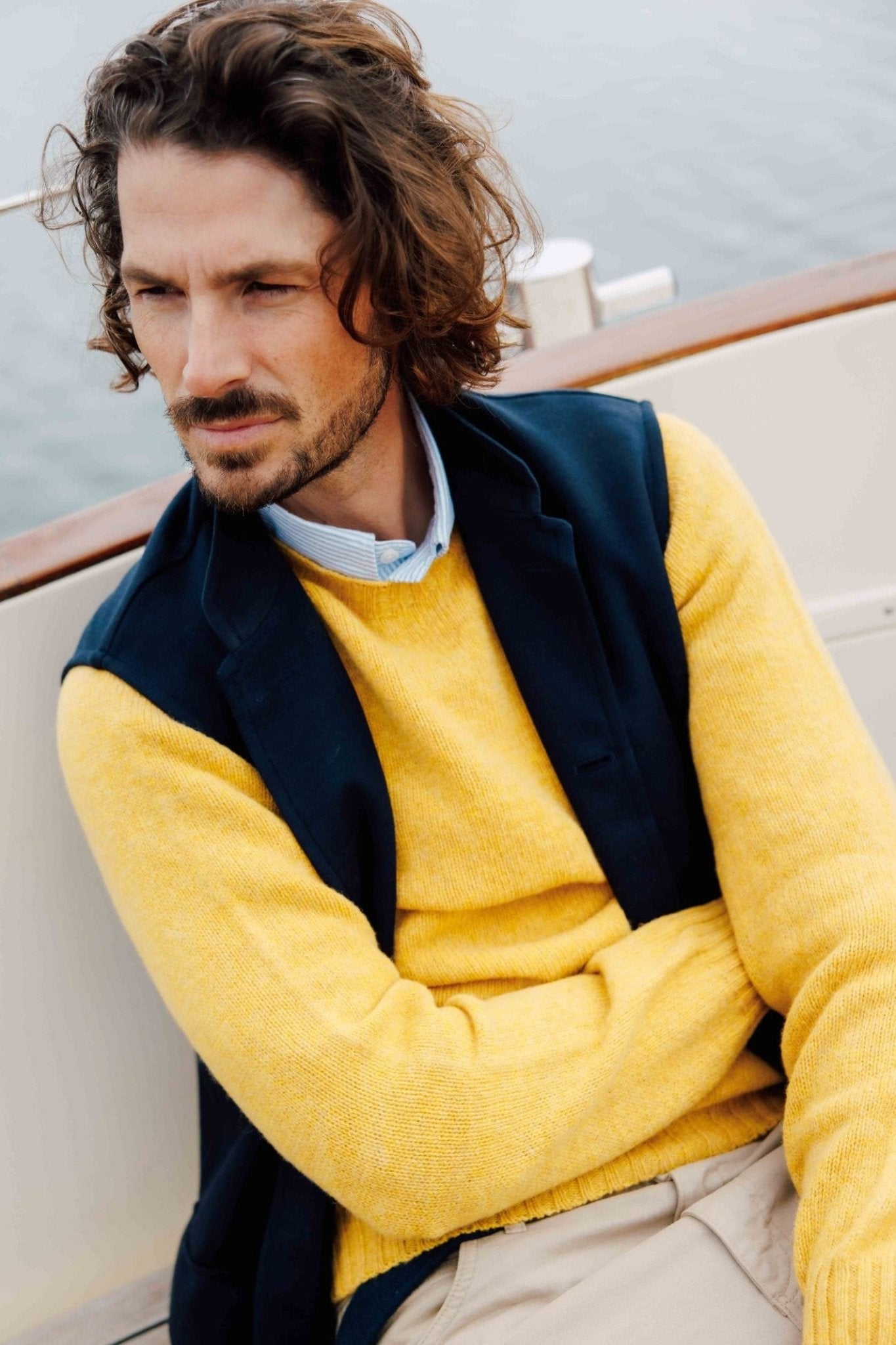 A man with wavy brown hair and a beard sits on a boat, wearing Campbells of Beauly Knitted Nehru Gilet over a blue shirt and yellow wool sweater, gazing thoughtfully to the side with water in the background.