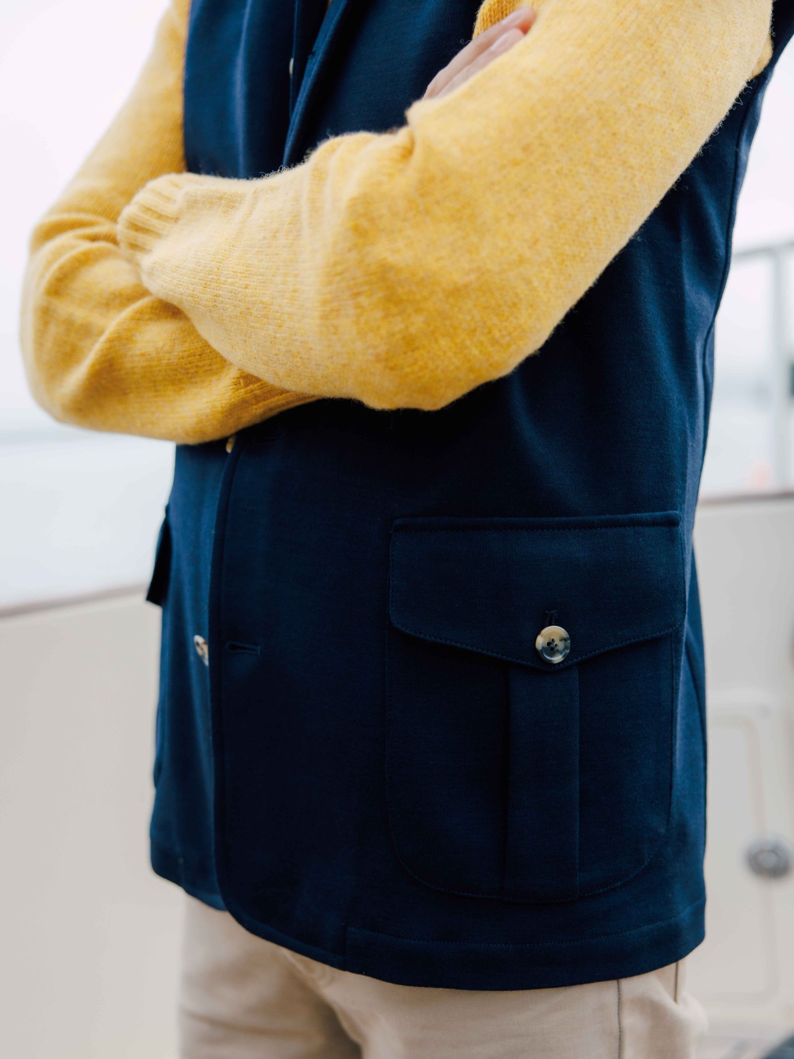 A person with crossed arms wears a yellow sweater under the Campbells of Beauly Knitted Nehru Gilet, made from 100% wool and featuring large front pockets, paired with beige pants—an ideal layering piece for outdoor or nautical settings.