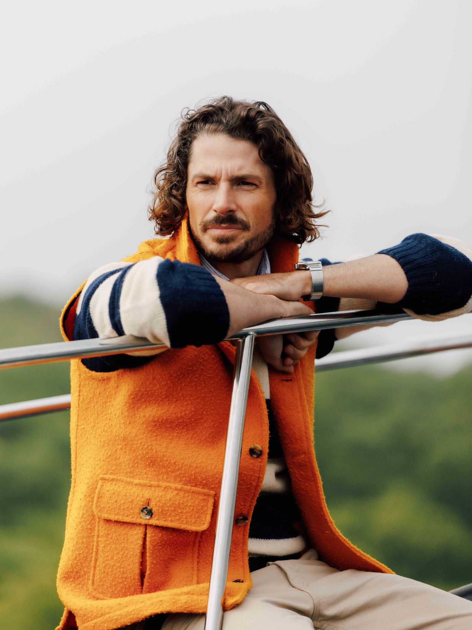 A man with curly brown hair and a beard leans on an outdoor metal railing, wearing a bright orange Campbells of Beauly Casetino Fleece Gilet over a navy and white striped sweater. Blurred green foliage appears in the background.