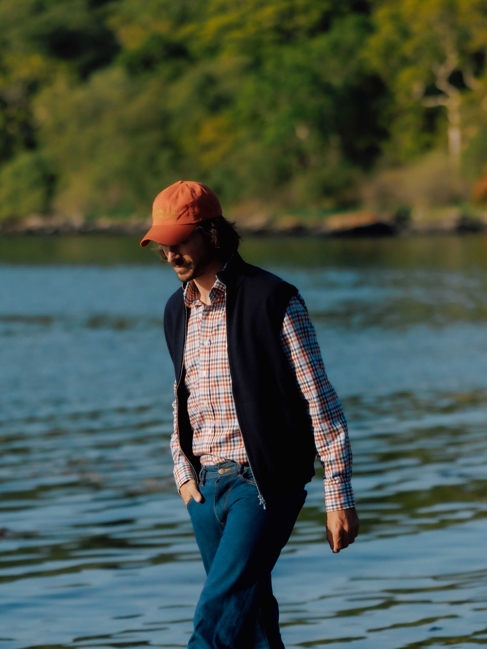 A person in a red cap, plaid shirt, and Campbells of Beauly Zip Gilet strolls beside a calm body of water, classic style highlighted against a backdrop of blurred green trees.