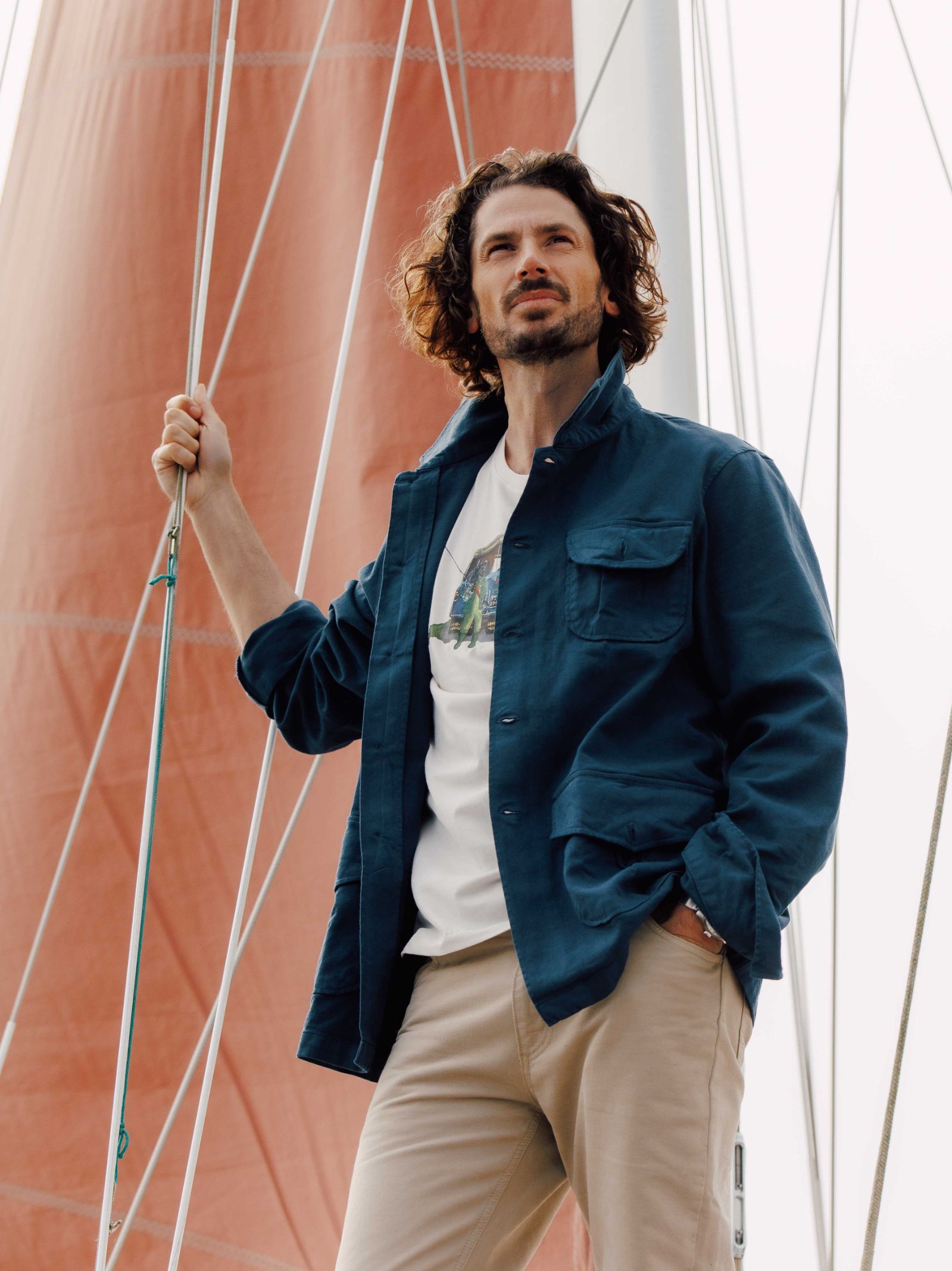 A man with wavy, shoulder-length hair stands on a sailboat, holding a rope. He wears Campbells of Beaulys Field Chore Jacket in blue over a white shirt and beige pants, with rigging and sail in the background for a utilitarian look.