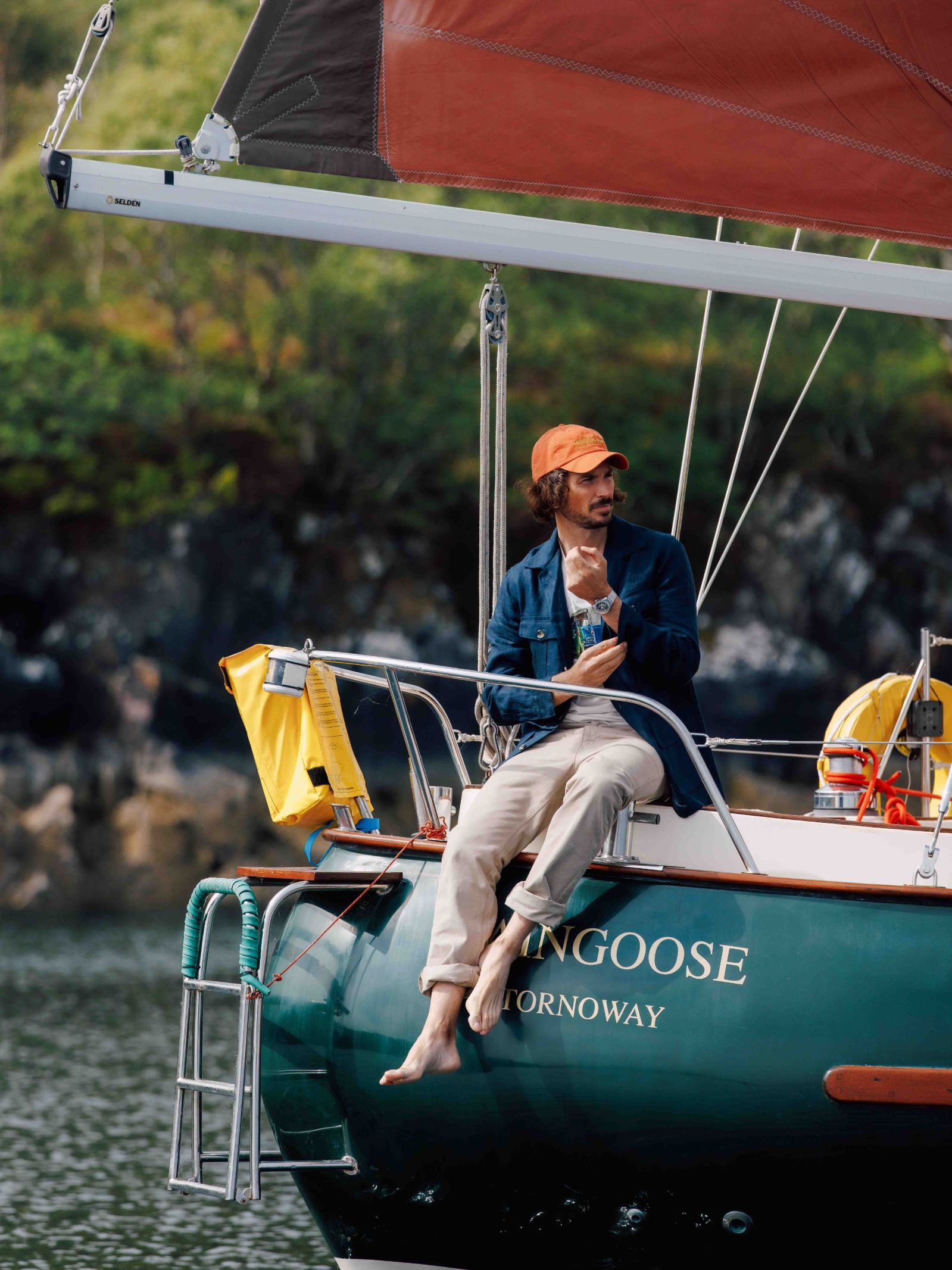 A man in a blue jacket, orange cap, and Campbells of Beauly Neutral Cotton Jeans sits barefoot on the edge of the green sailboat Mongoose Stornoway, drink in hand, with trees and rocky shore in the background.
