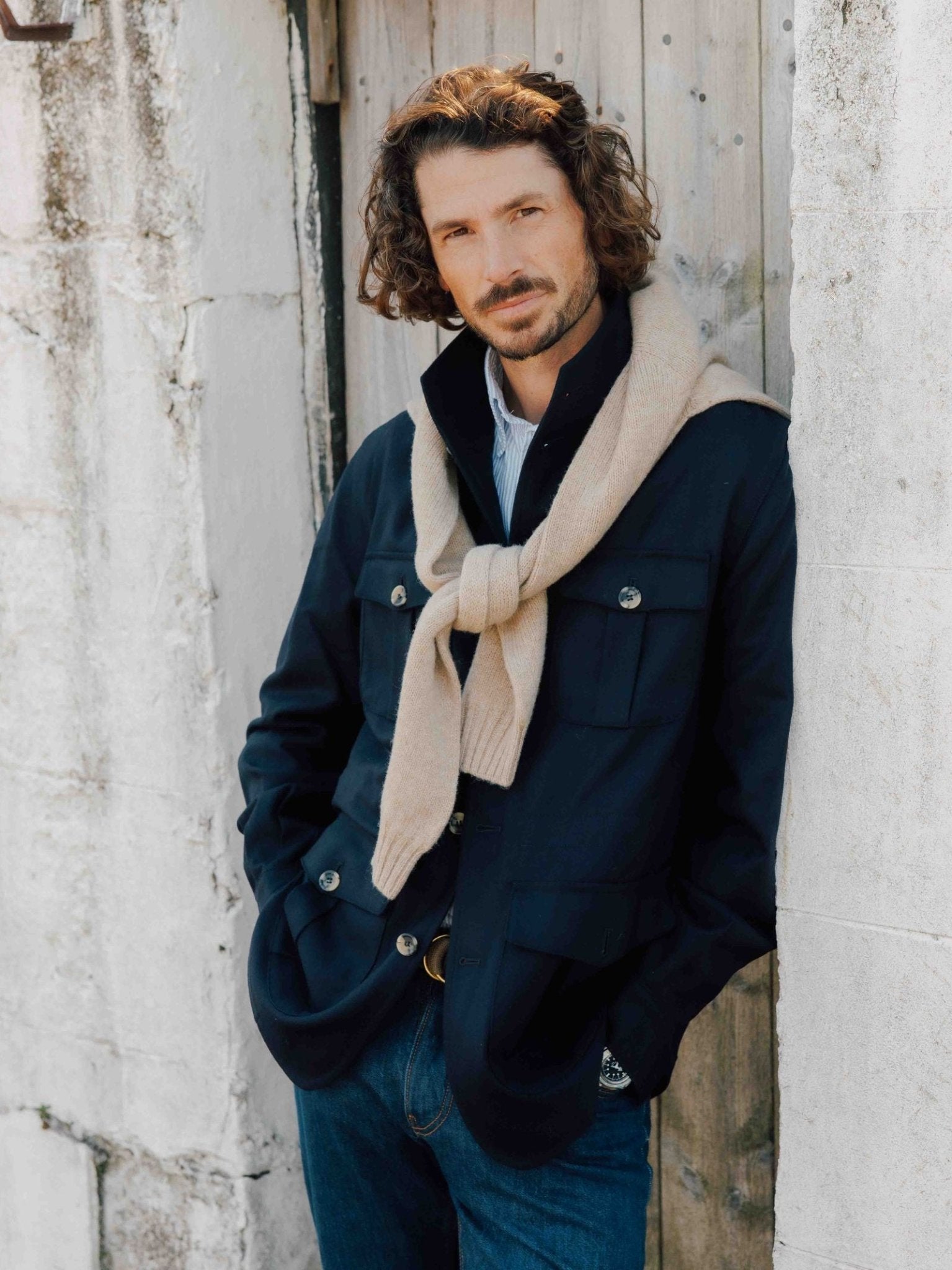 A man with wavy brown hair stands against a rustic wooden door, wearing Campbells of Beauly Field Teba Jacket, blue jeans, and a light beige sweater over his shoulders. He has his hands in his pockets and looks calmly at the camera.