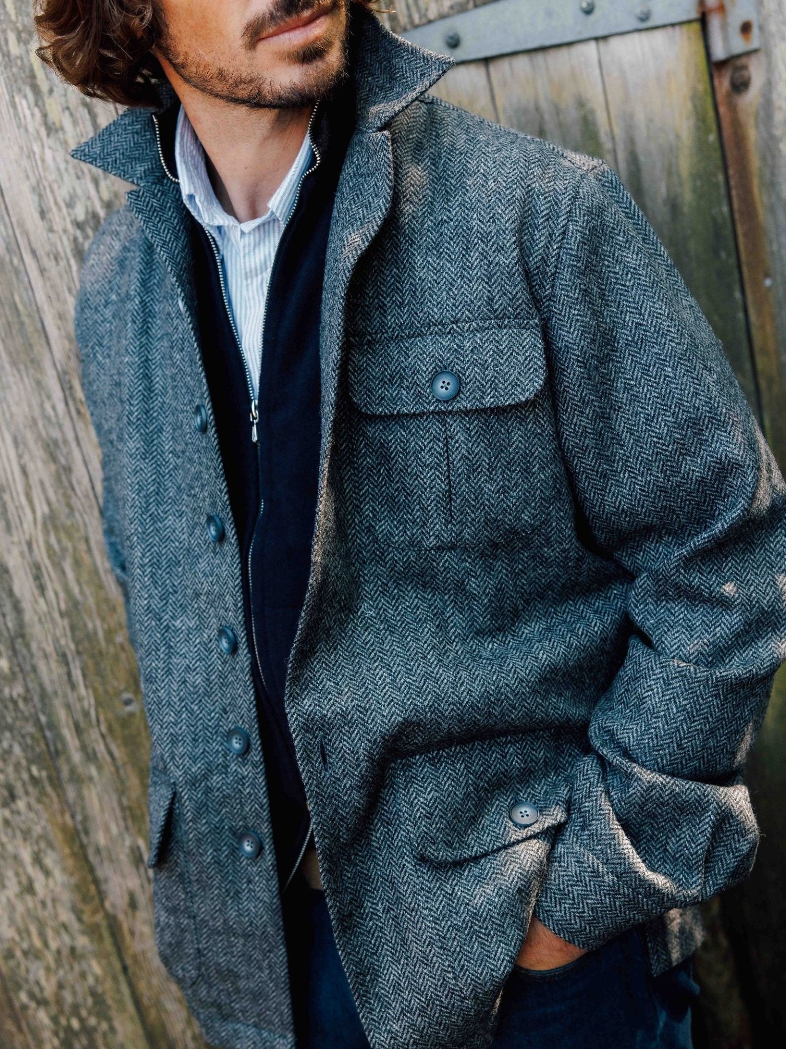 A man wearing the Campbell's of Beauly Casual Shacket with flap pockets stands before a weathered wooden door, one hand in his pocket, looking to the side—effortlessly channeling relaxed tailoring.
