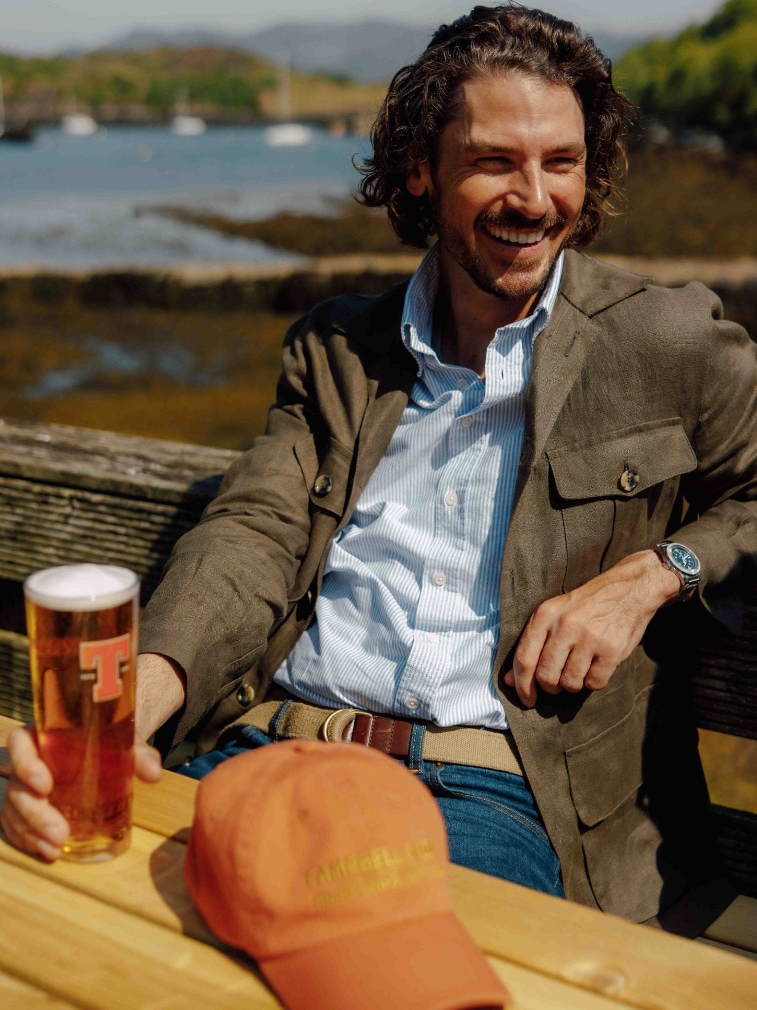 A smiling man with wavy hair sits outdoors by the water, wearing a Campbells of Beauly Linen Safari Jacket over his blue shirt and jeans. He holds a pint of beer at a wooden table, an orange cap nearby and hills in the background.