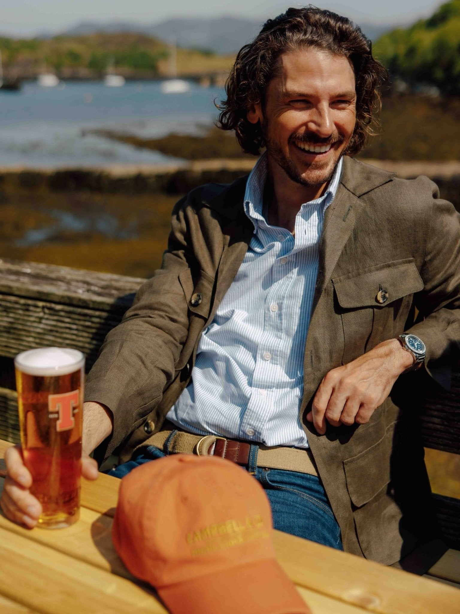 A smiling man with wavy brown hair sits at an outdoor wooden table by the water, wearing a Campbell’s of Beauly Oxford Shirt, a brown blazer, and jeans. An orange cap with yellow embroidery rests on the table beside his amber beer.
