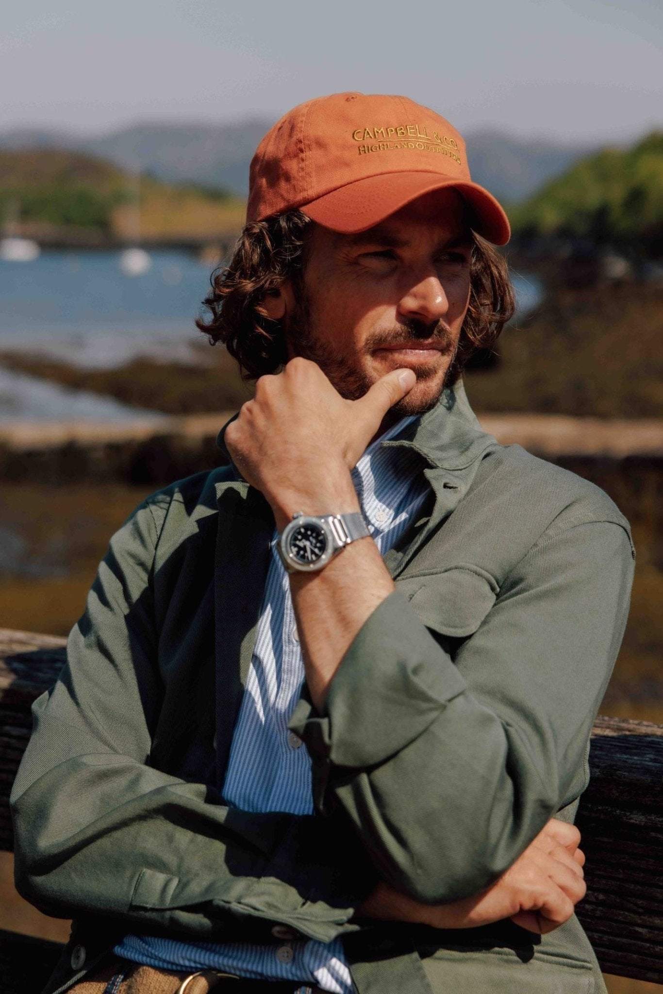 Wearing the Campbells Baseball Cap by Campbells of Beauly, a man with wavy hair and a beard sits on a wooden bench by the water, embodying comfort and style as he gazes thoughtfully to the side.
